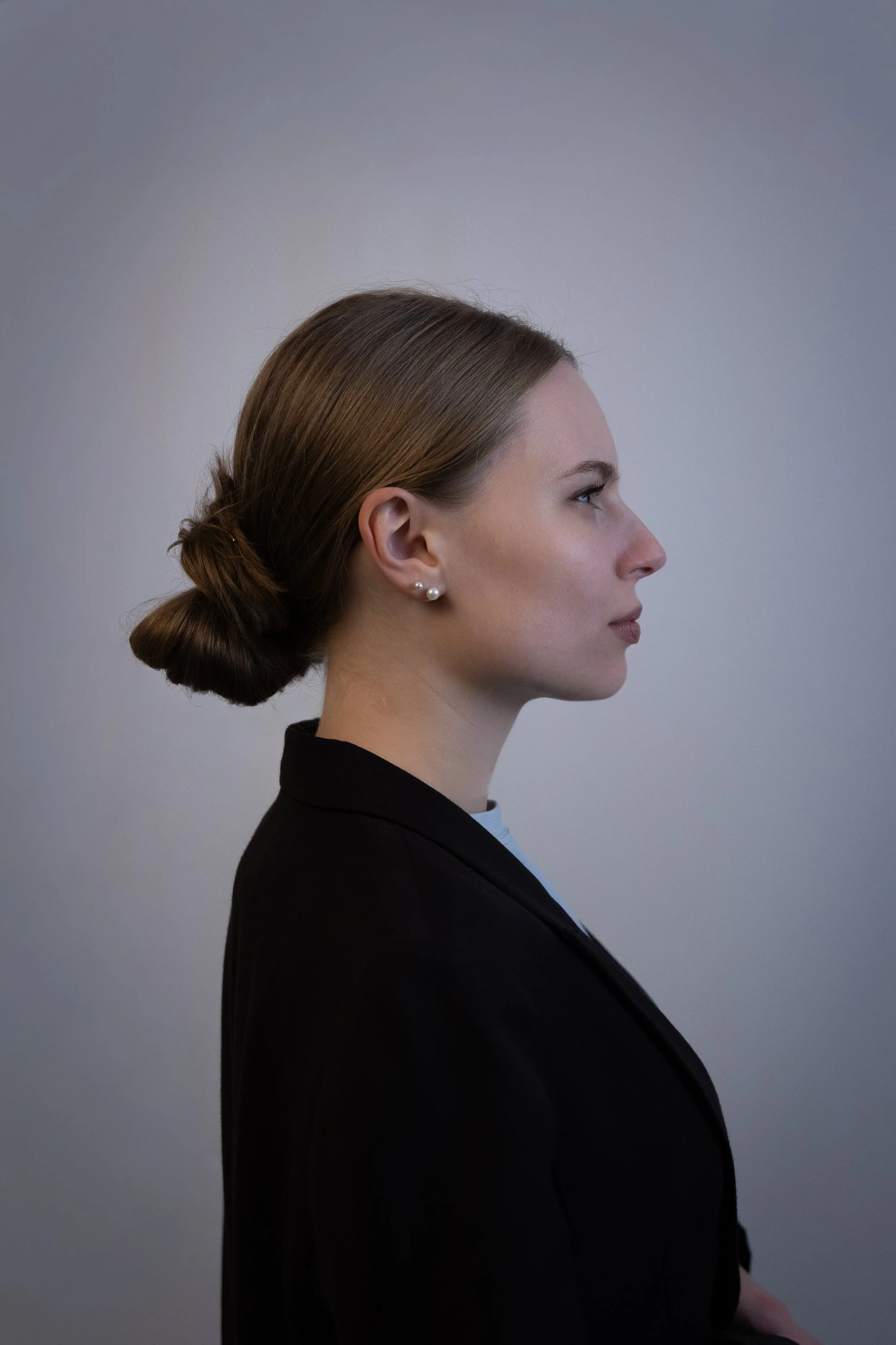 Seitenprofil einer Frau mit braunen Haaren, hoher Hochsteckfrisur, Perlenohrringen, in schwarzem Blazer und weißem Hemd, vor einem neutralen Hintergrund.