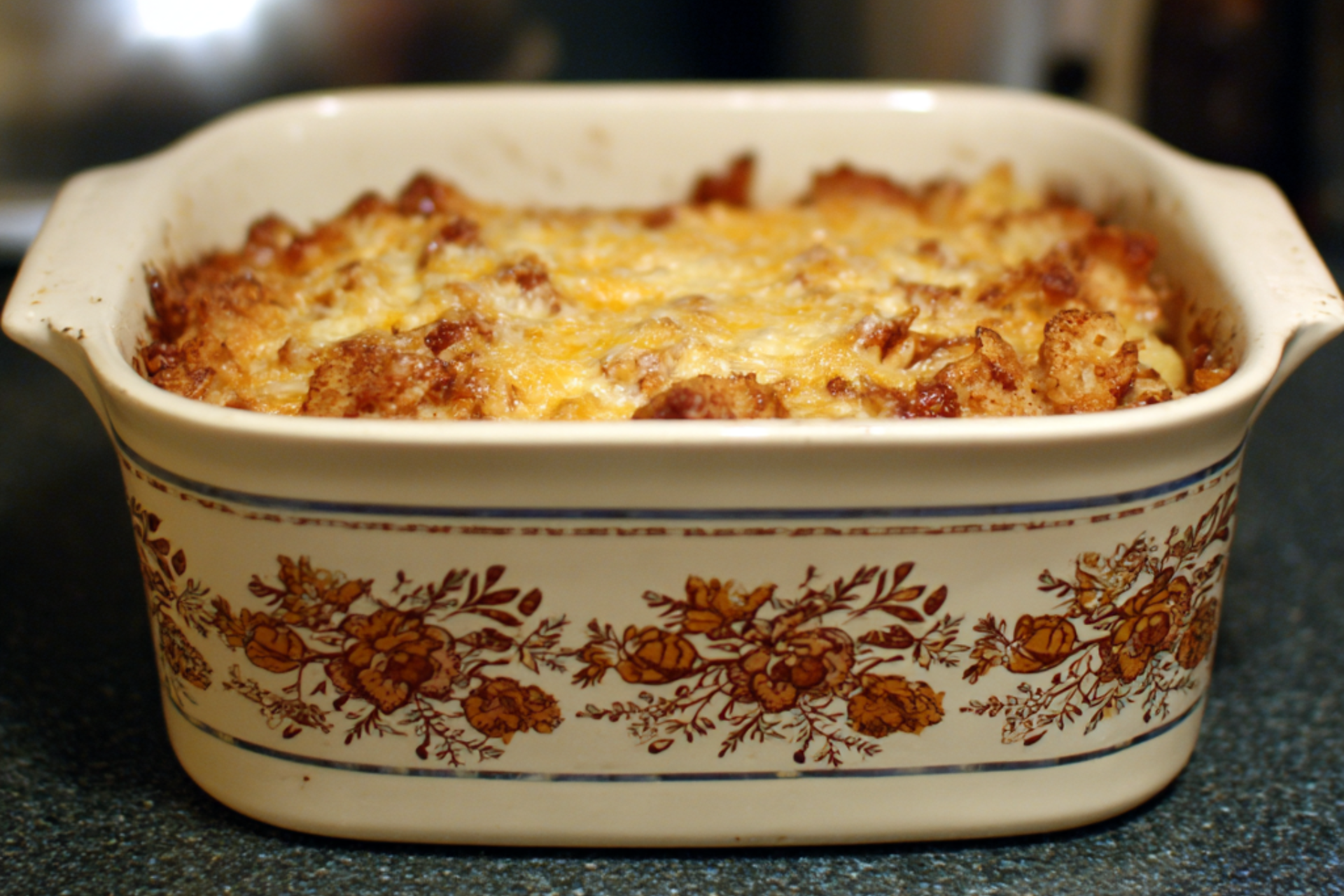 A baked casserole in a vintage floral ceramic dish resting on a kitchen counter, with a browned, slightly uneven top.