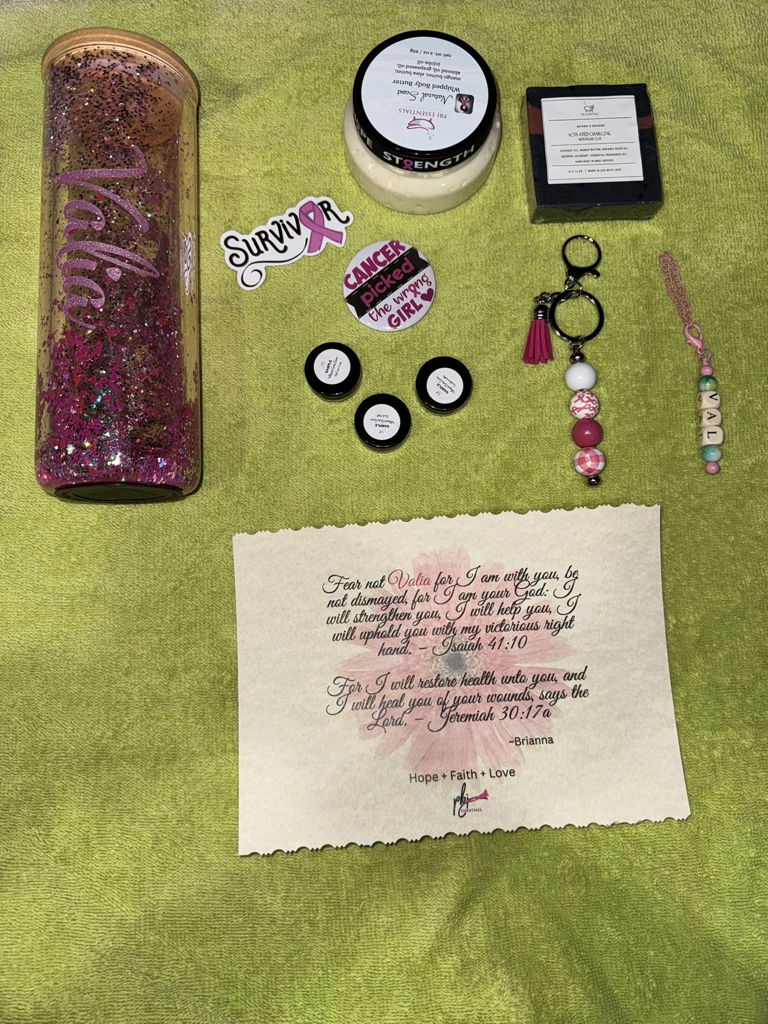 Collection of items including a pink glittered water bottle, skincare products, a ring, beaded jewelry, and motivational stickers on a green cloth.