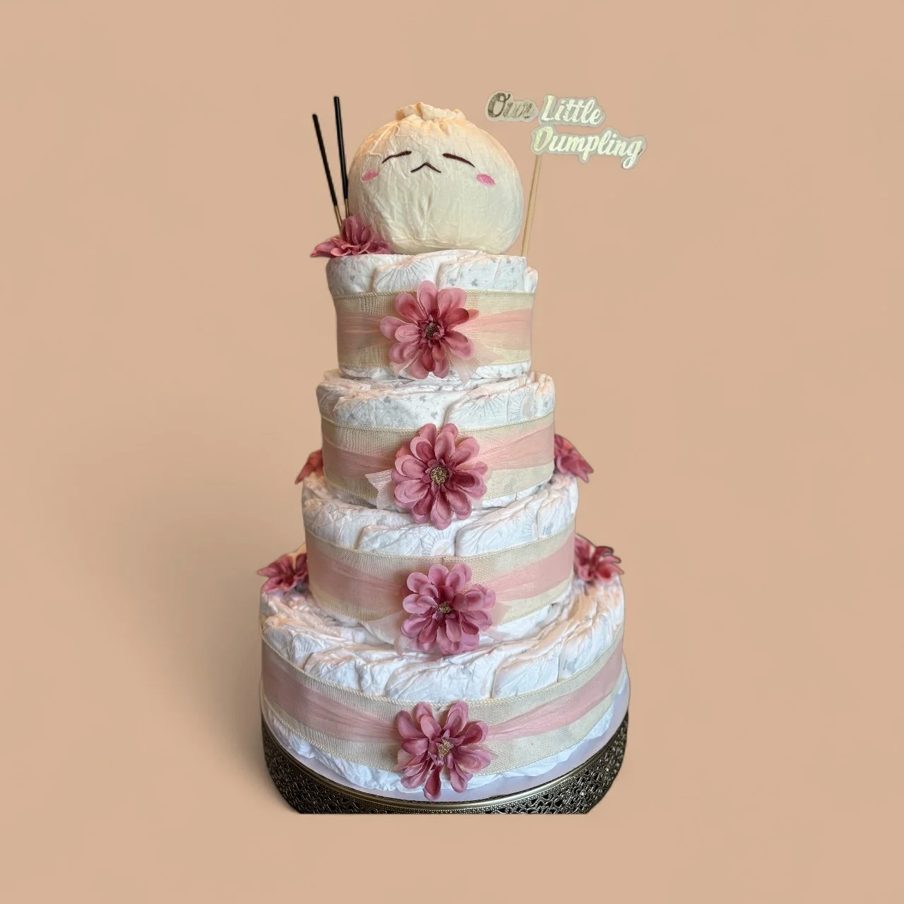 A tiered diaper cake decorated with pink flowers and pastel ribbons, topped with a plush dumpling figure holding chopsticks and a sign that reads 'Our Little Dumpling'.