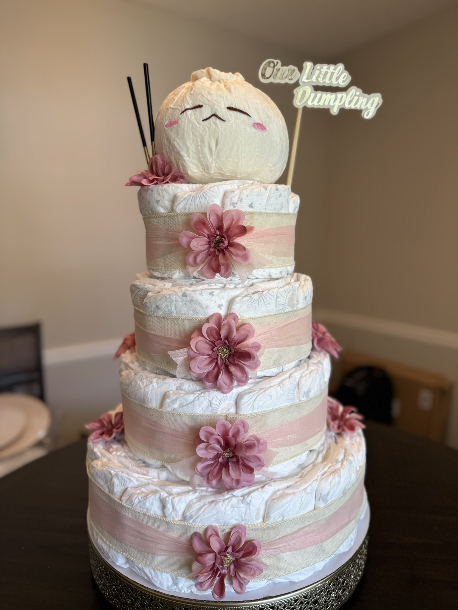 A diaper cake decorated with pink flowers and ribbons, topped with a plush dumpling character with a happy face and a sign that reads 'One Little Dumpling'.