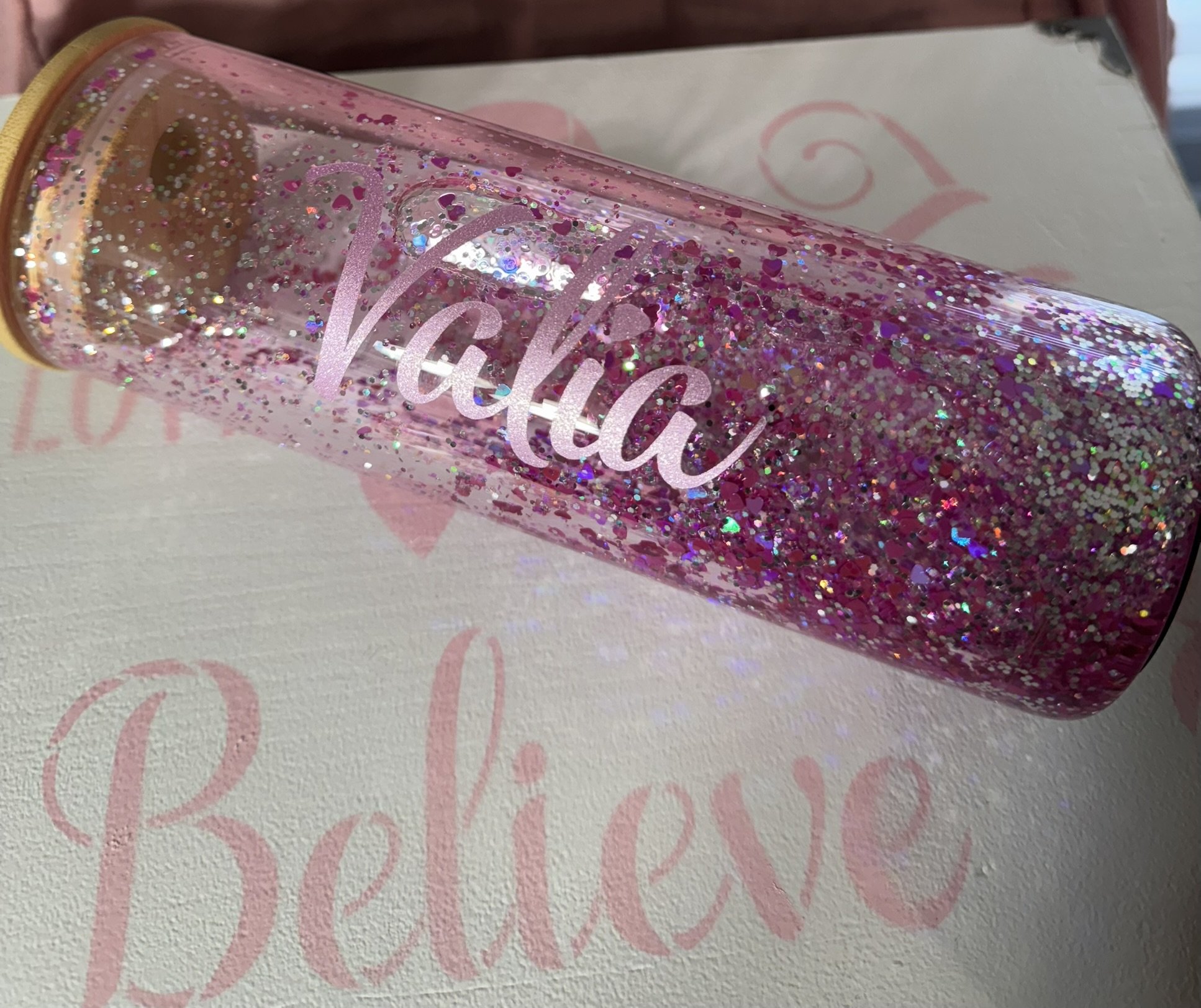 Pink glittery water bottle with the word 'Valeria' written on it in cursive, placed on a surface with the word 'Believe' faintly visible in pink.