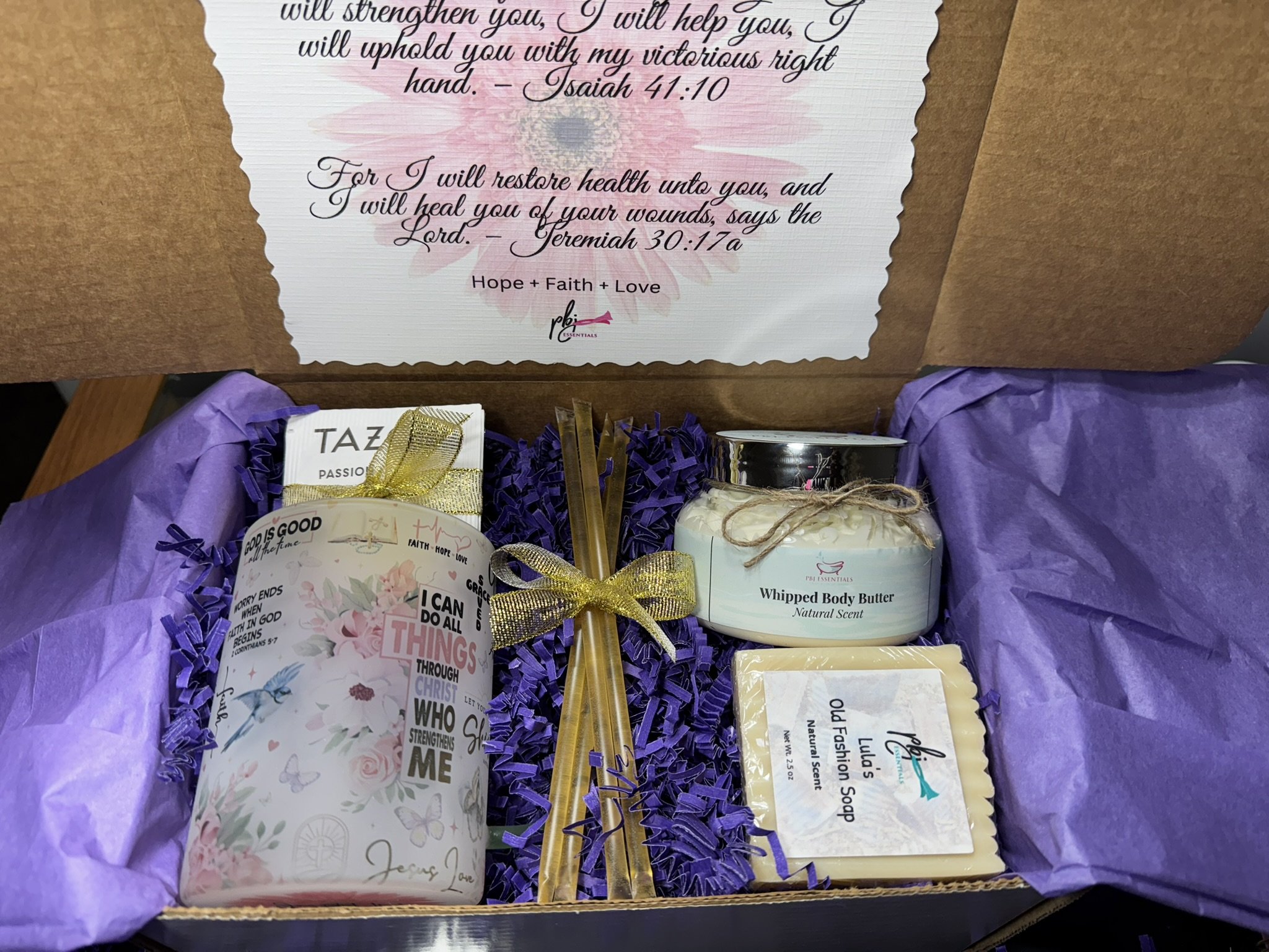 Open gift box with purple tissue paper filled with various items including a candle, a jar of whipped body butter, a bar of soap, and a tea mug. The box has a handwritten-style message on the inner lid with Bible quotes and words of encouragement.