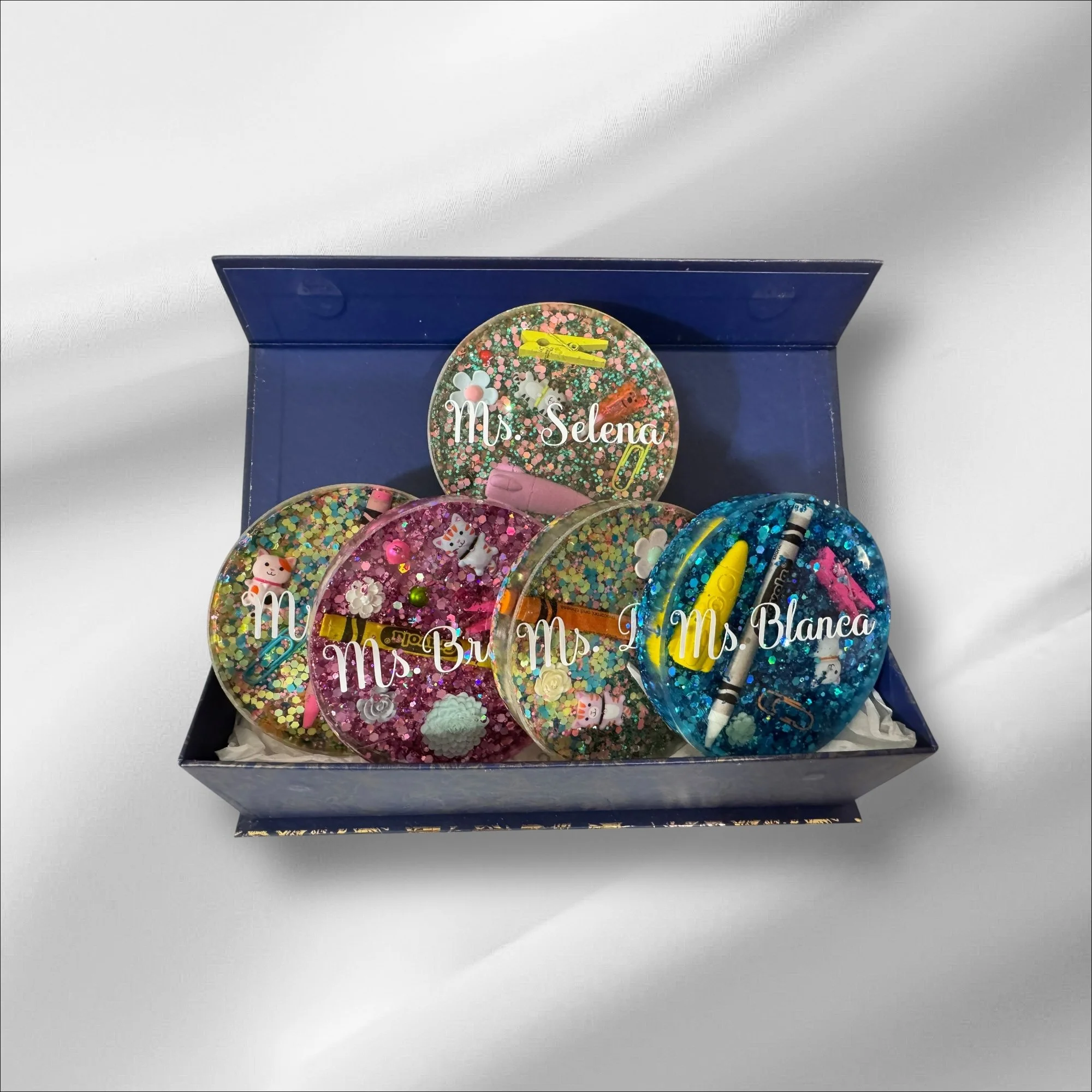Decorative resin coasters with glitter, miniature toys, and charms inside, labeled with personal names, in a dark blue box on a white surface.