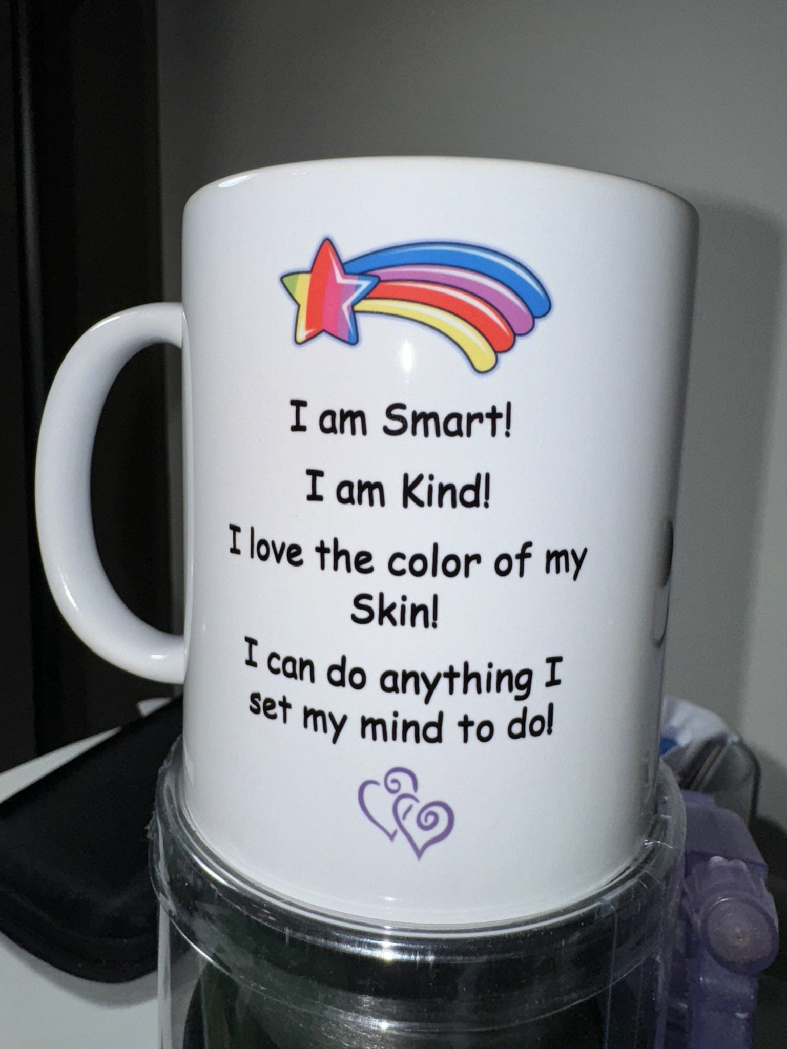 White mug with colorful star and rainbow design, featuring positive affirmations about intelligence, kindness, skin color, and confidence, placed on a clear plastic cup or container.