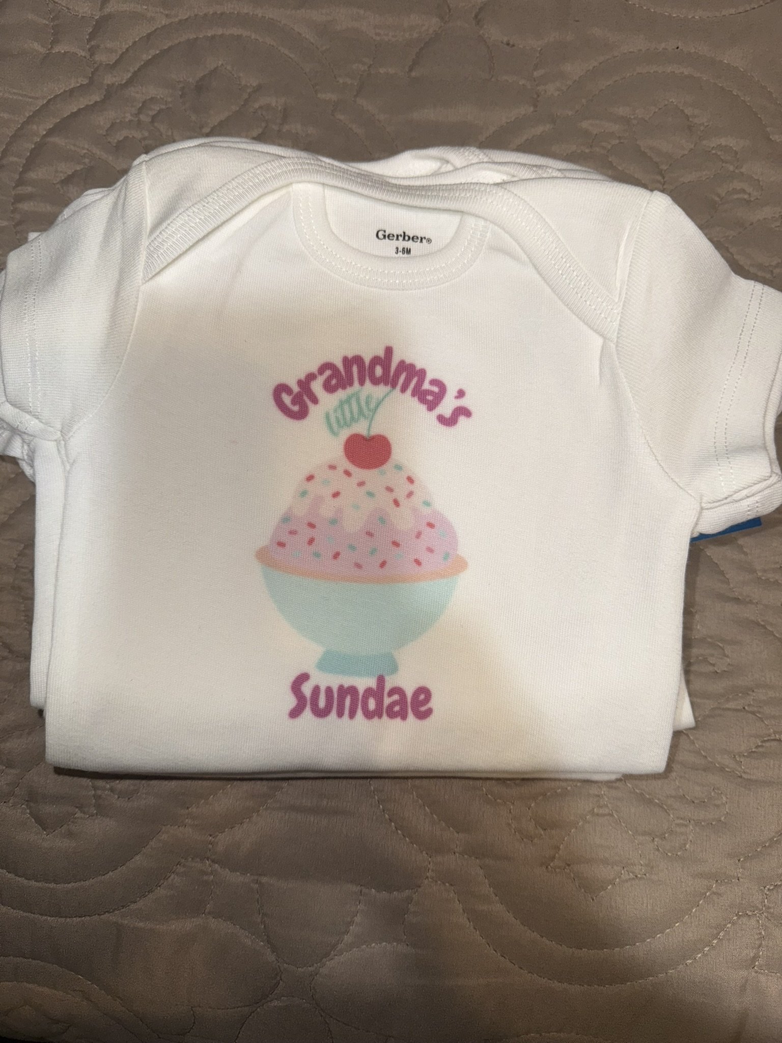 A white infant's onesie with a printed design featuring a bowl of pink ice cream with sprinkles, a cherry on top, and the text "Grandma's July Sundae" in colorful letters.