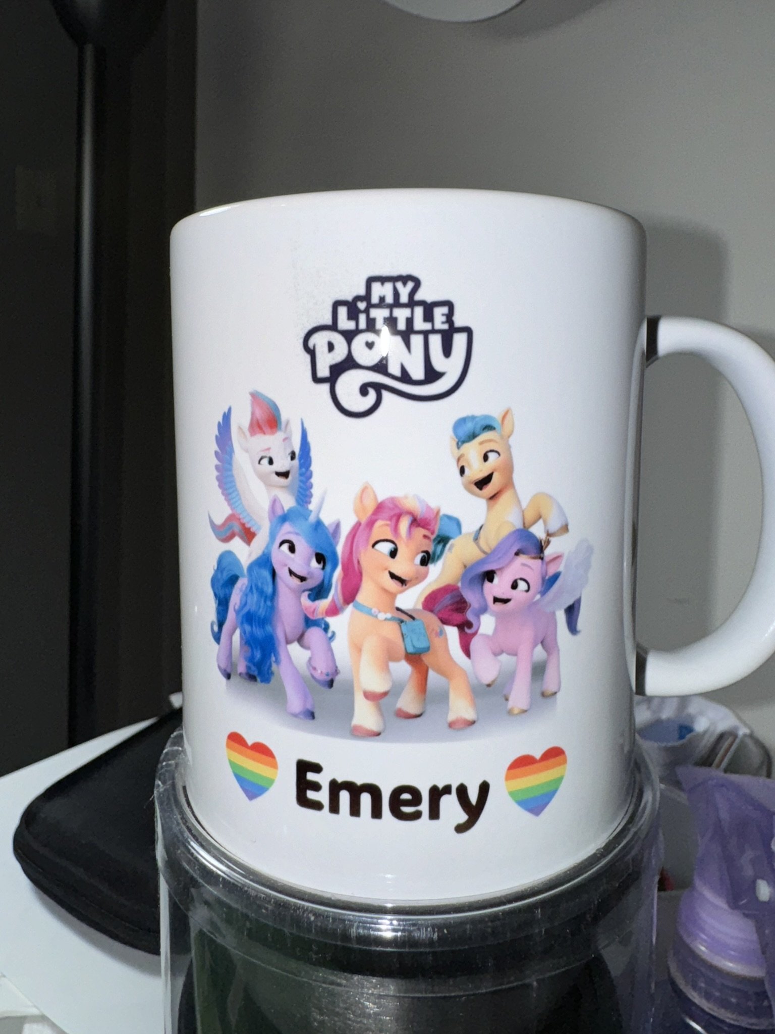 A white ceramic mug with 'My Little Pony' characters and the name 'Emery' with rainbow heart graphics.