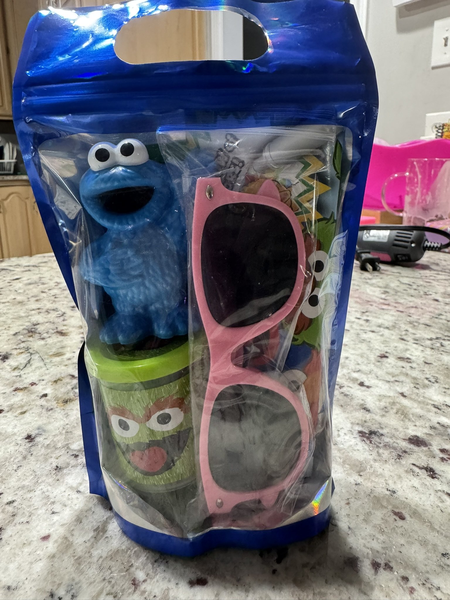 A blue Sesame Street Cookie Monster toy, pink sunglasses, and cup with Sesame Street character faces inside a sealed plastic bag on a kitchen countertop.