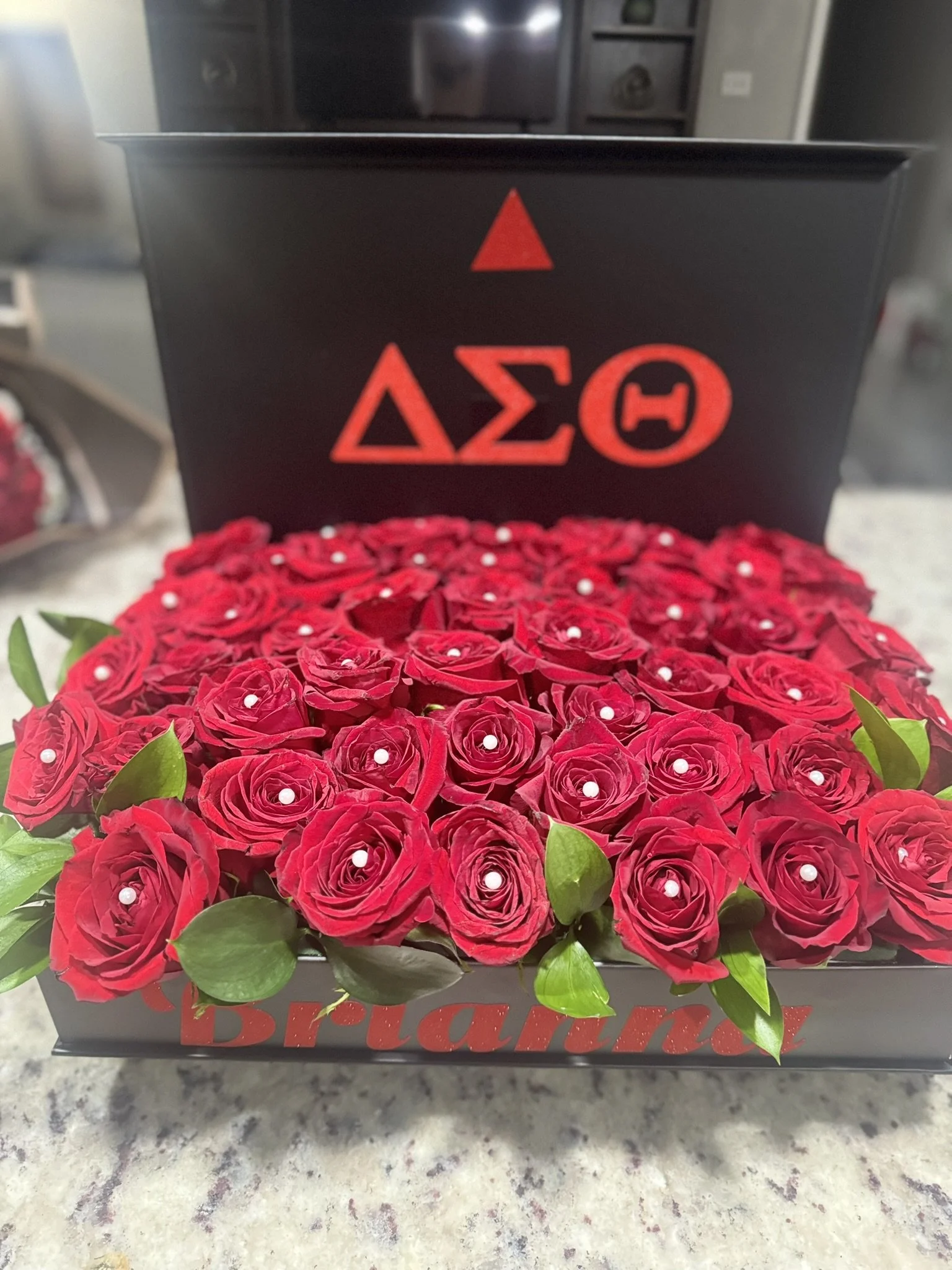 A black box with the Greek letters Delta Sigma Theta and a red triangle and circle symbol on the lid. Inside the box, there are red roses decorated with small white pearls, with some green leaves visible at the edges.