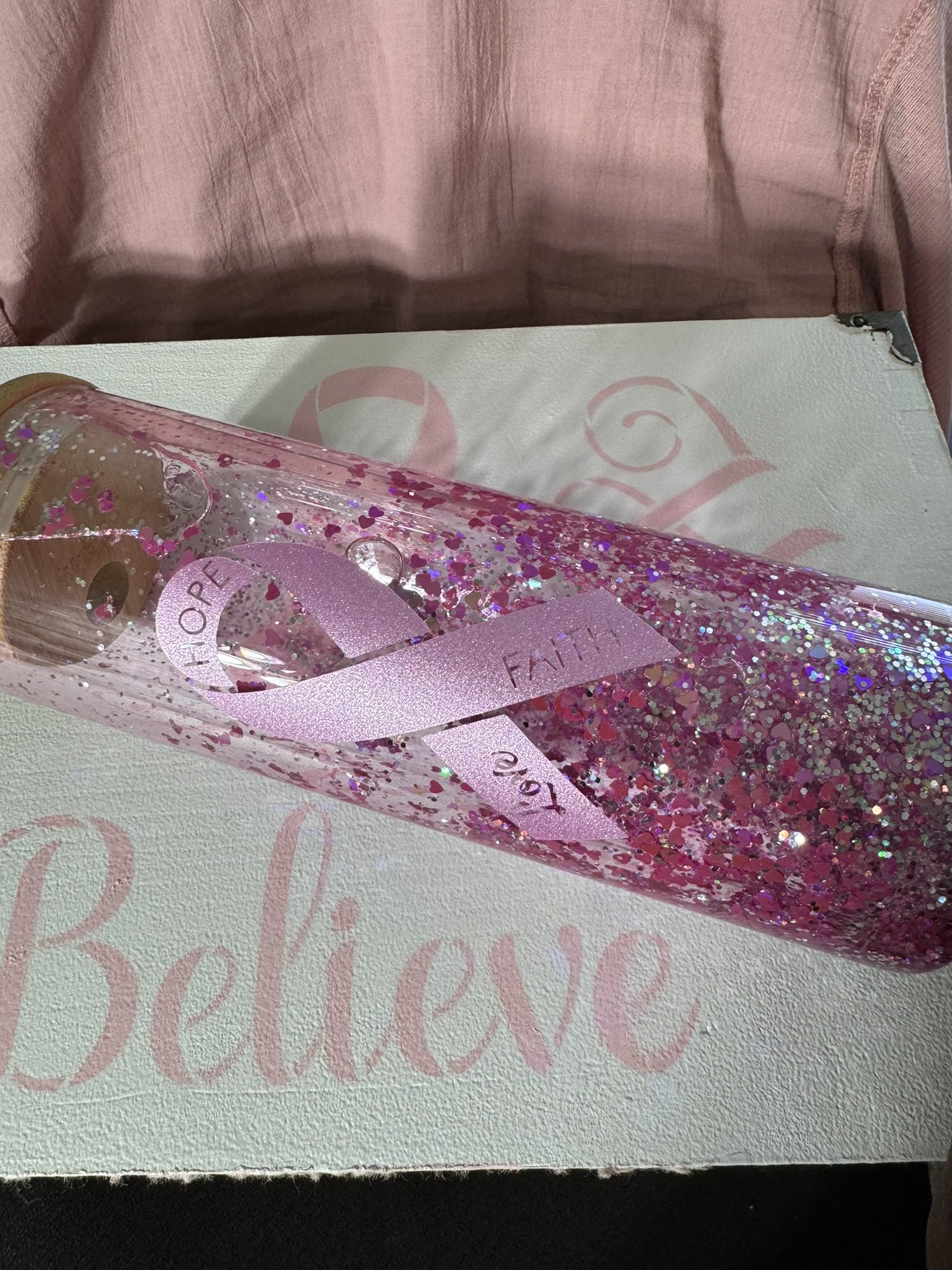 A pink glittery bottle with the words 'Hope', 'Faith', and 'Love' written on it, resting on a box with the word 'Believe' printed on it, with a pink cloth in the background.