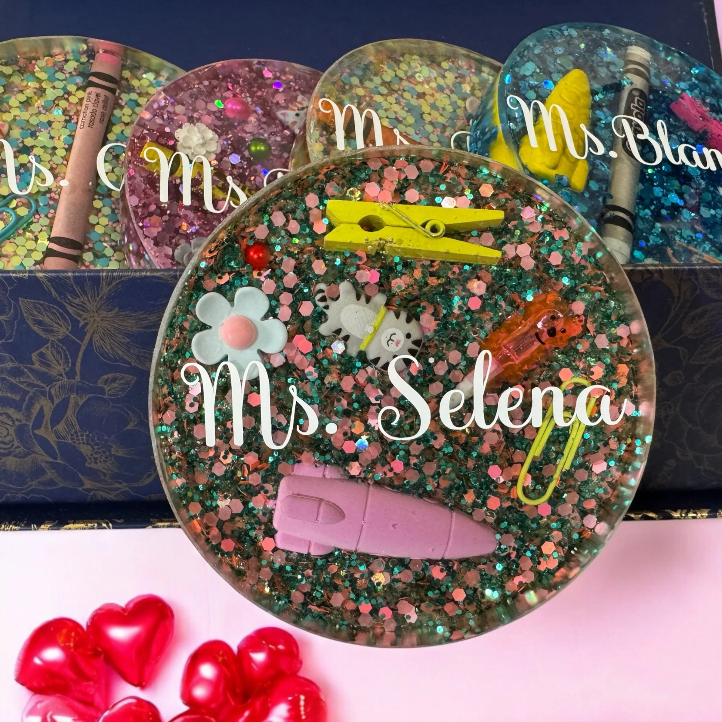 A glittery resin coaster with small decorative items including a white flower, a pink donut, a yellow clothespin, a small orange cartoon character, and a small sheep sticker, with the text 'Ms. Selena' written across it.