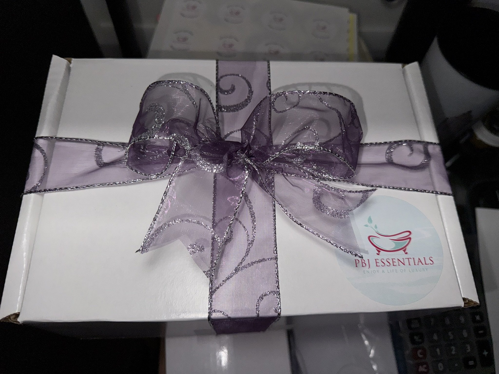 A white gift box wrapped with a lavender ribbon bow that has a glittery swirl design. The box has a logo sticker from PBJ Essentials, featuring a pink bathtub graphic and the tagline 'Enjoy a Life of Luxury'.