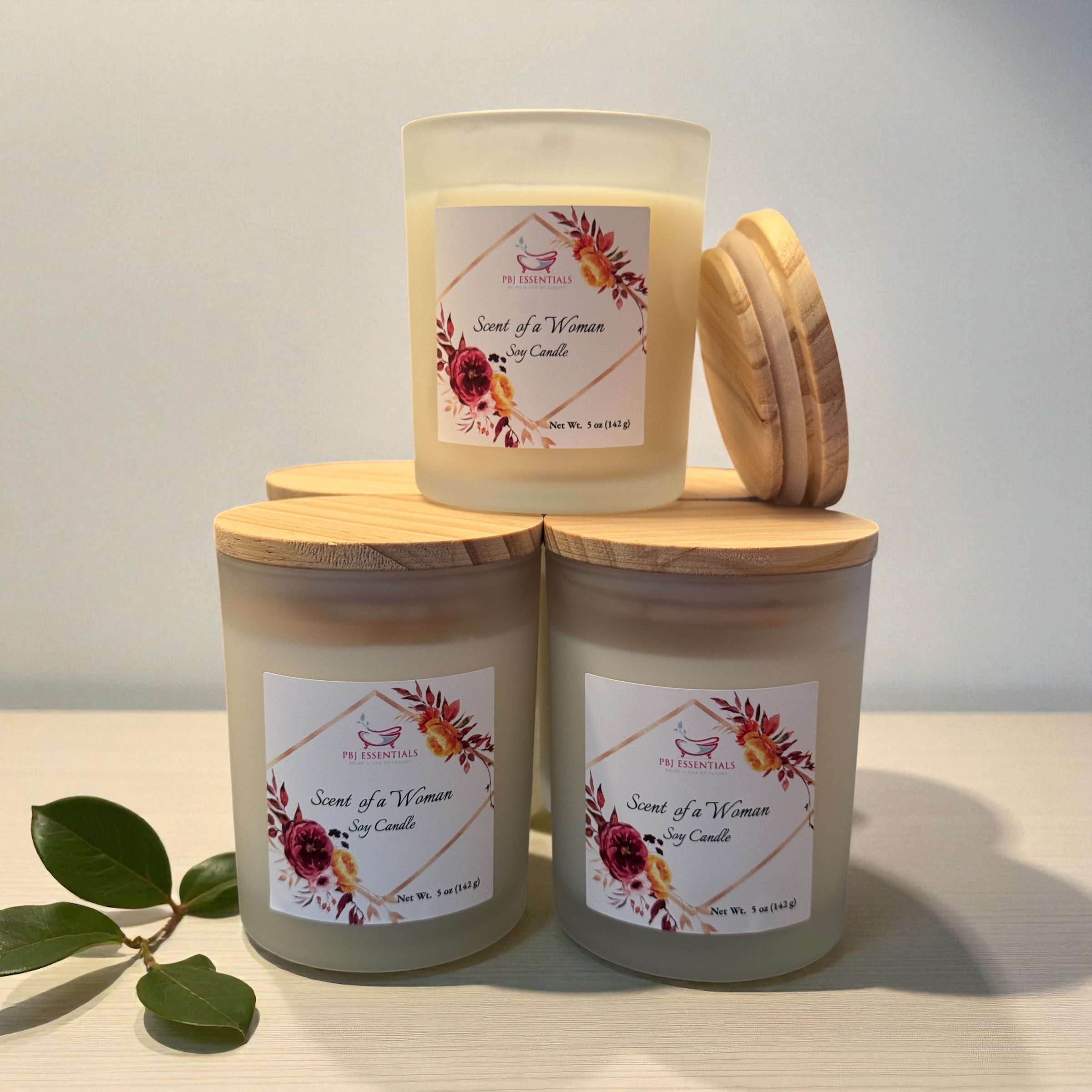Three scented soy candles labeled "Scent of a Woman" by PBJ Essentials, with wooden lids, arranged in a pyramid on a light-colored surface, with green leaves nearby.