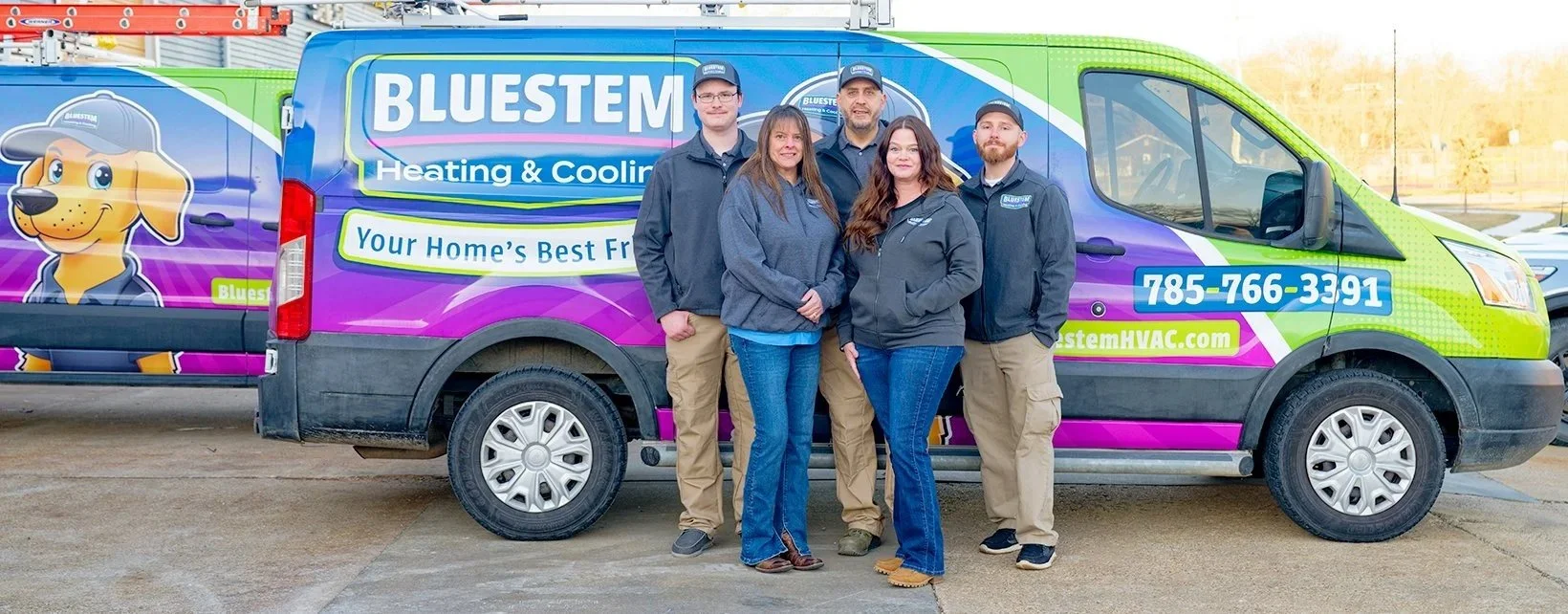 Four people standing in front of a colorful service van for Bluestem Heating & Cooling, with a cartoon dog mascot and contact information displayed.