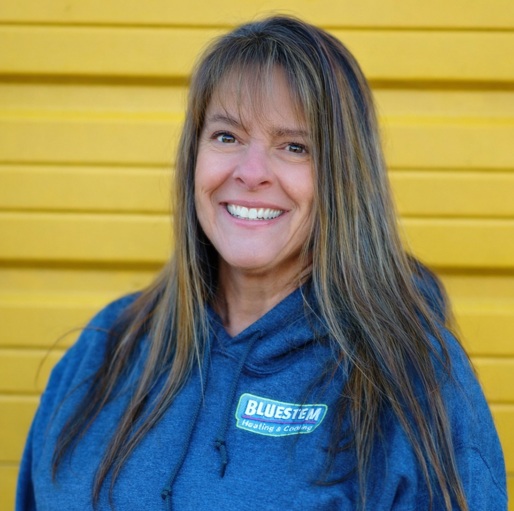Debbie Knudsen, a woman smiling in front of a yellow wall, wearing a blue hooded sweatshirt with a 'BLUESTEM Heating & Cooling' logo.