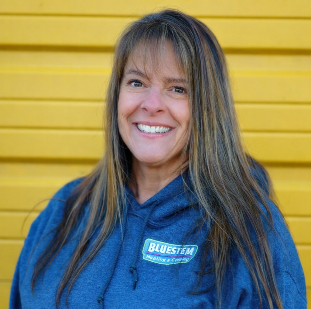 A woman with long brown hair and a big smile, wearing a blue hoodie with a logo that says "BLUESTEM Heating & Cooling," standing in front of a bright yellow wall.