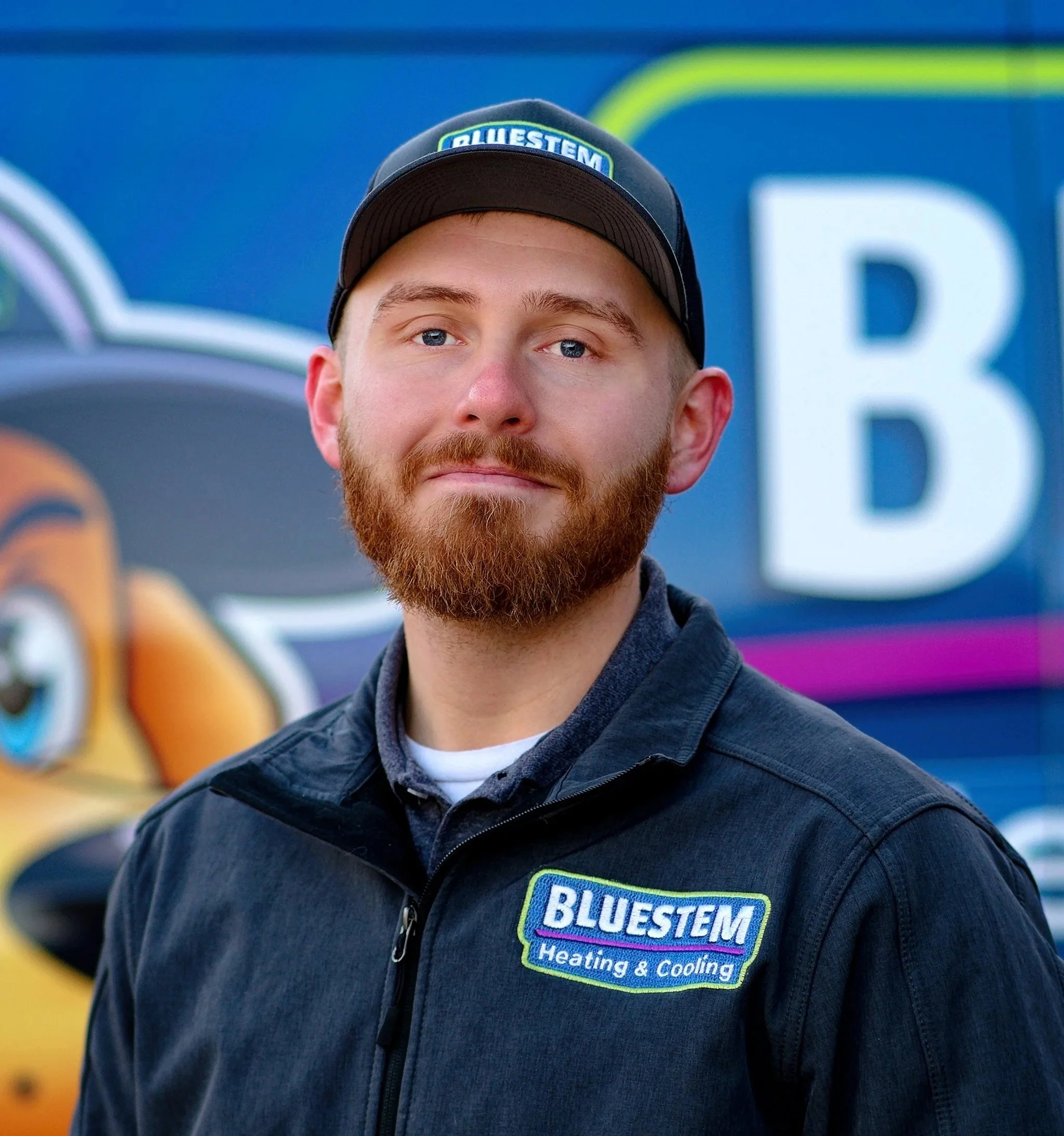 A young man with a beard and blue eyes wearing a black jacket with 'BLUESTEM Heating & Cooling' logo, standing outside in front of a blue background with a large letter 'B' and part of a cartoon lion illustration.