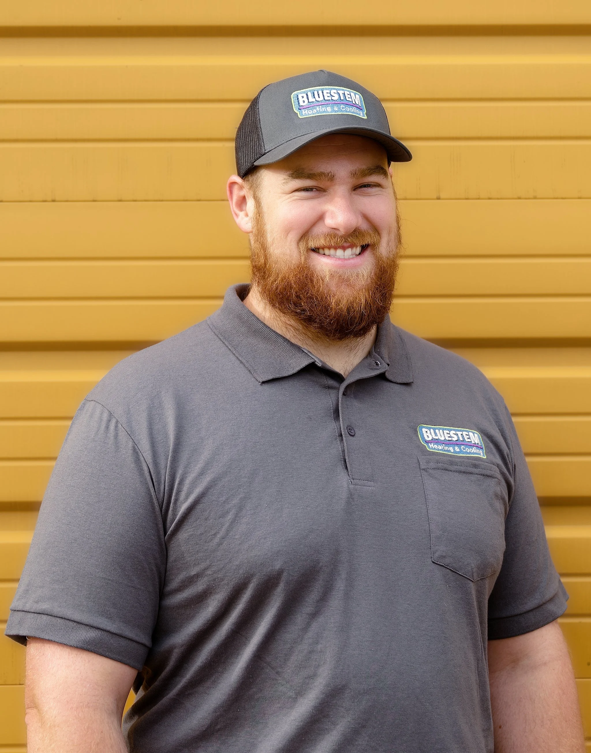 A man with a beard and light skin wearing a gray polo shirt and a gray baseball cap with a 'Blustem Heating & Cooling' logo, standing in front of a yellow horizontal slat wall, smiling.