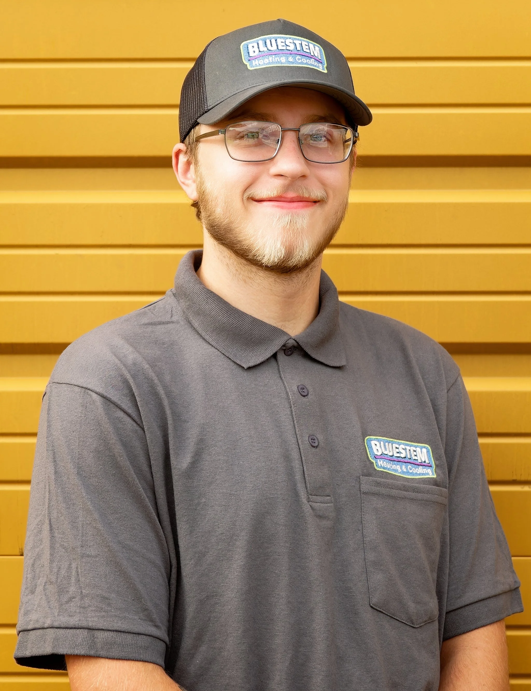 A young man with glasses and a beard wearing a gray polo shirt and a gray baseball cap with a logo for Bluestem Heating & Cooling, standing in front of a yellow wooden wall.