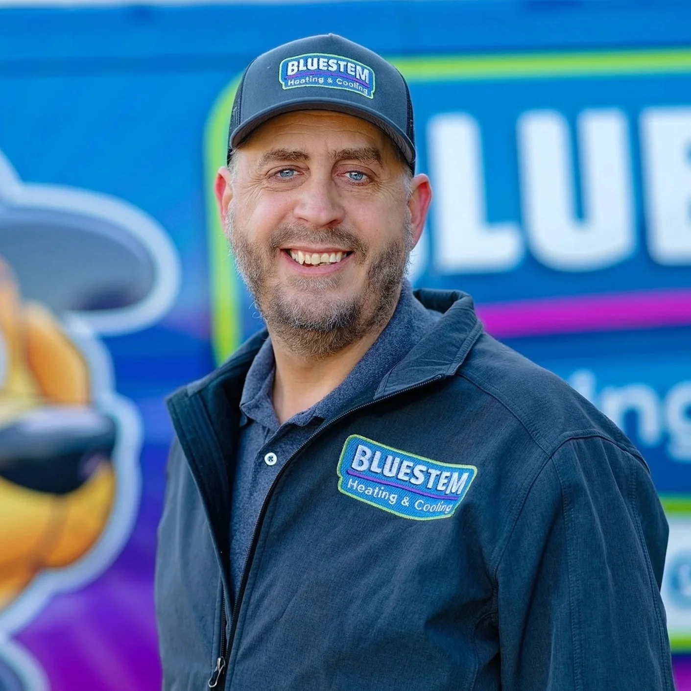 Jason Fisher, a man in a dark gray jacket and cap smiling, with a logo that reads "BLUESTEM Heating & Cooling," standing outdoors in front of a blue and purple Bluestem promotional truck.