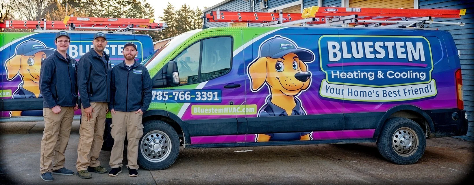 Three men standing in front of a company van with colorful branding for Bluestem Heating & Cooling. The van features a cartoon dog mascot wearing a cap, and has ladders attached to the roof. The men are dressed in navy jackets and khaki pants.
