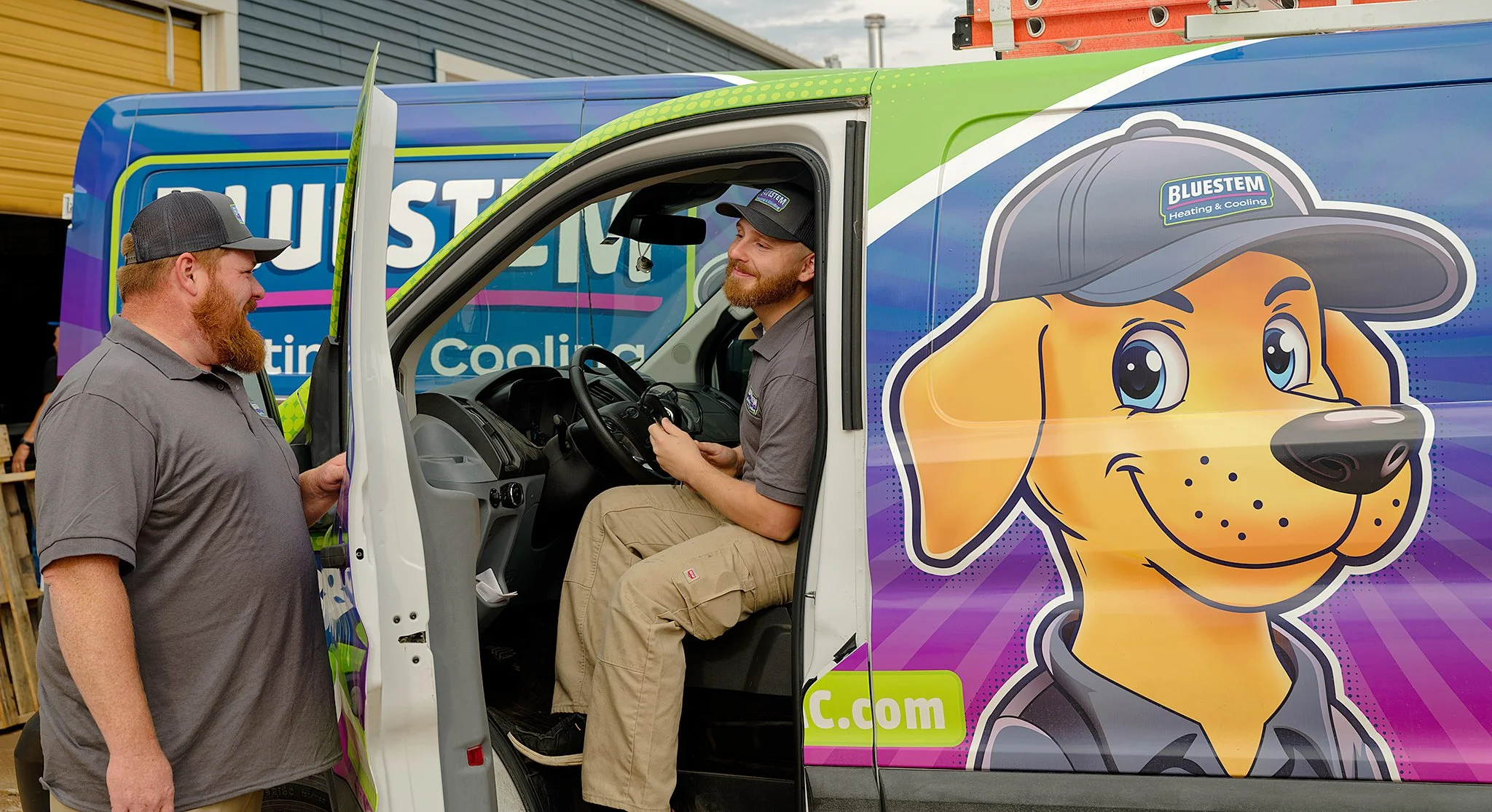 Two men in gray shirts and black caps talking near a Bluestem Heating & Cooling branded service van with cartoon dog graphics, one sitting inside the van and the other standing outside.