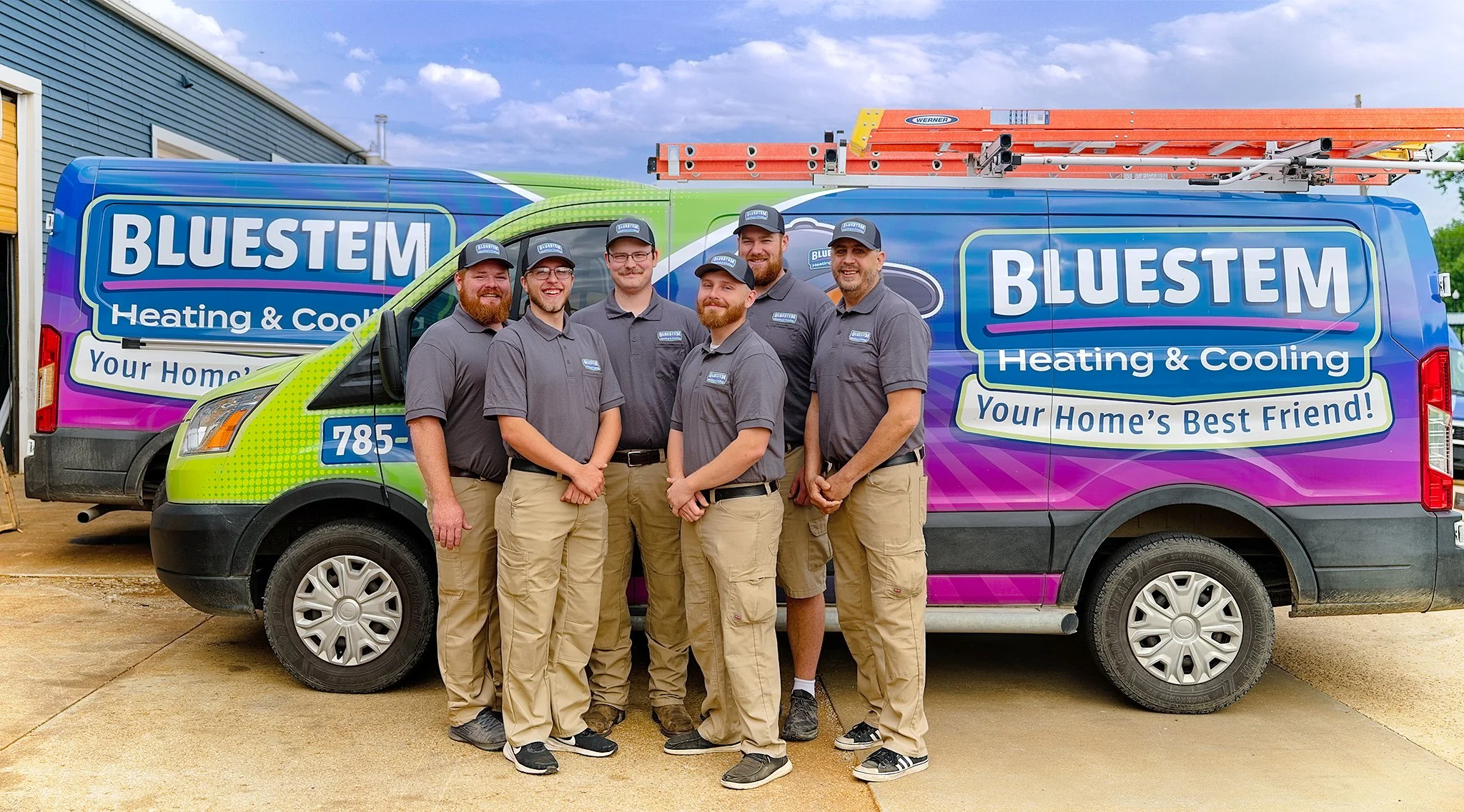 Group of six men in gray uniforms and black hats standing in front of a colorful van with 'BLUESTEM Heating & Cooling' branding, parked outdoors on a concrete surface with a house and blue sky in the background.