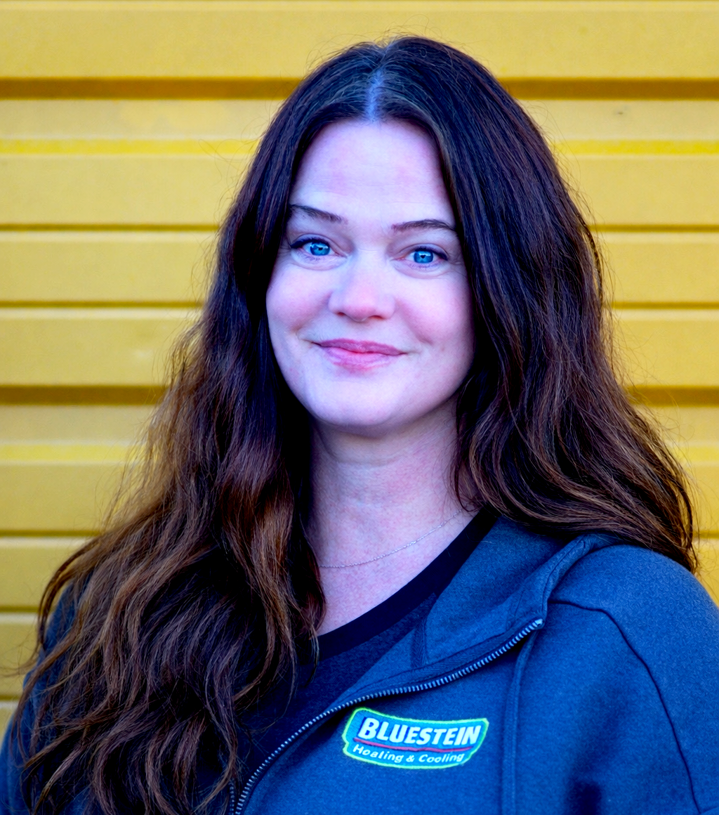 Rebecca McAnulla, a woman with long brown hair and blue eyes is smiling at the camera, wearing a dark gray jacket with a "Bluestem" logo, standing against a yellow metal background.