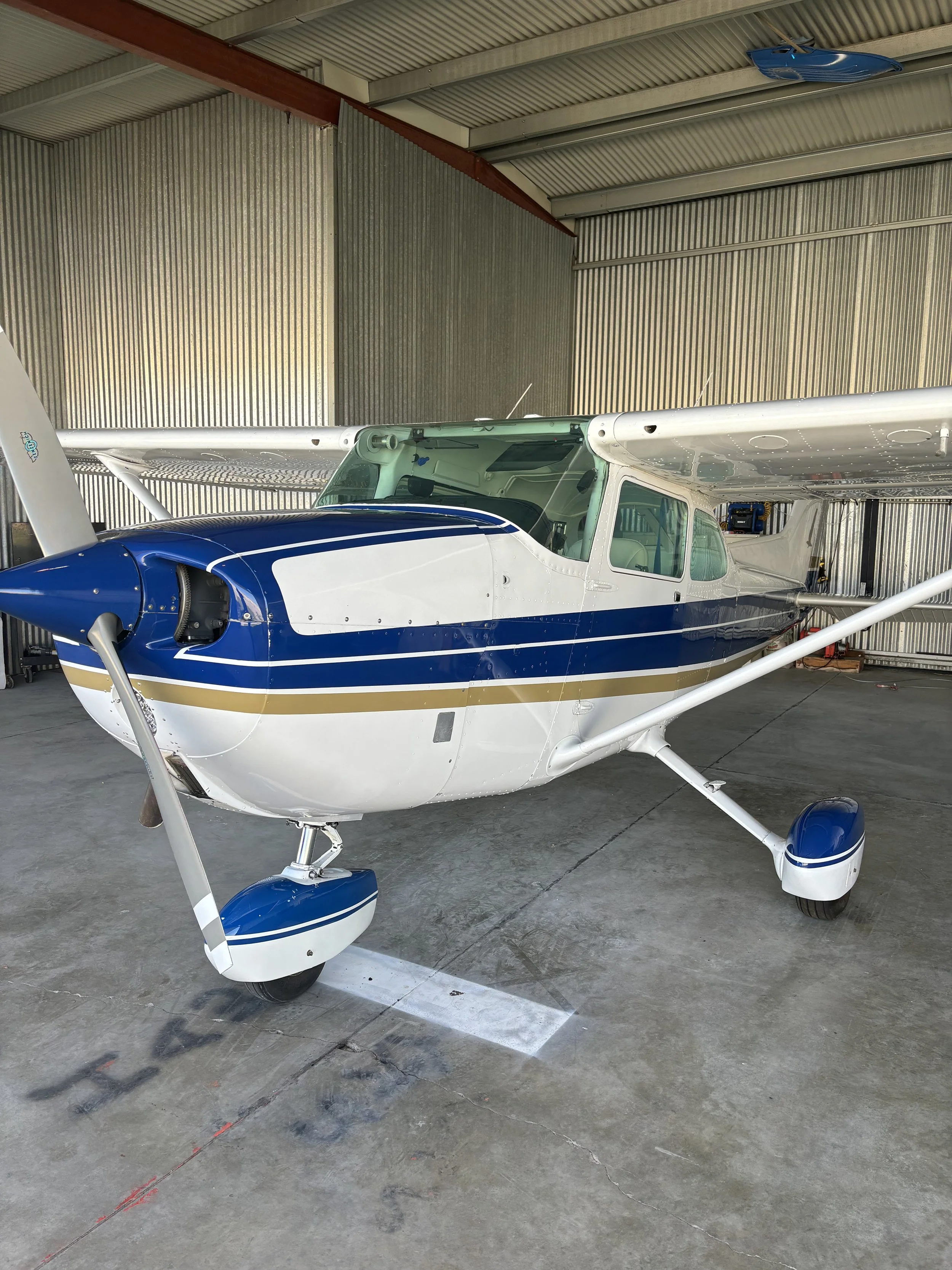 Modern rectangular building with wood and metal exterior, large glass window, set against mountainous landscape with dry grass and small trees in foreground.