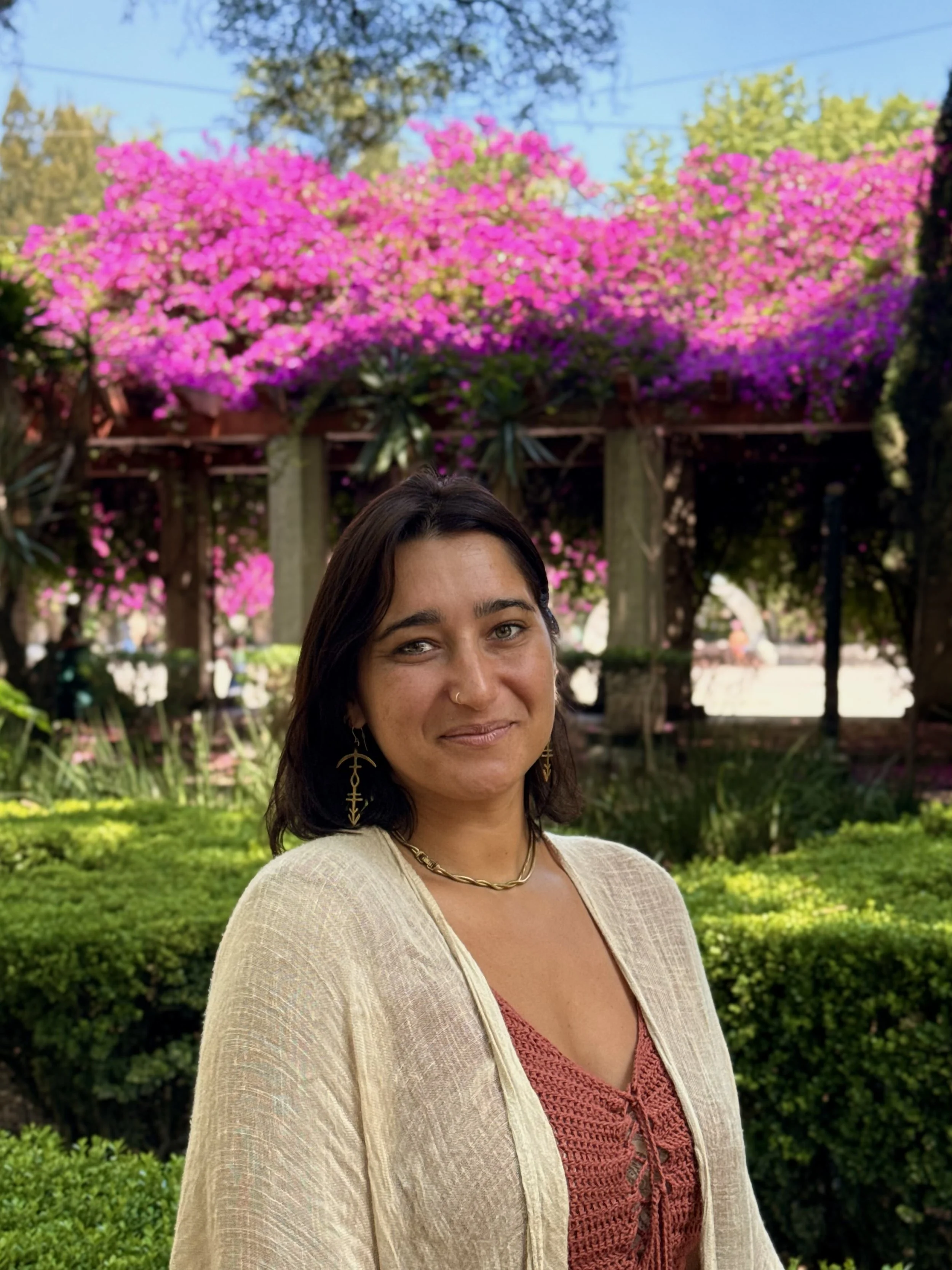 A woman with dark hair, wearing earrings, a necklace, a beige cardigan, and a red patterned top, standing outdoors in front of pink flowering trees and greenery.