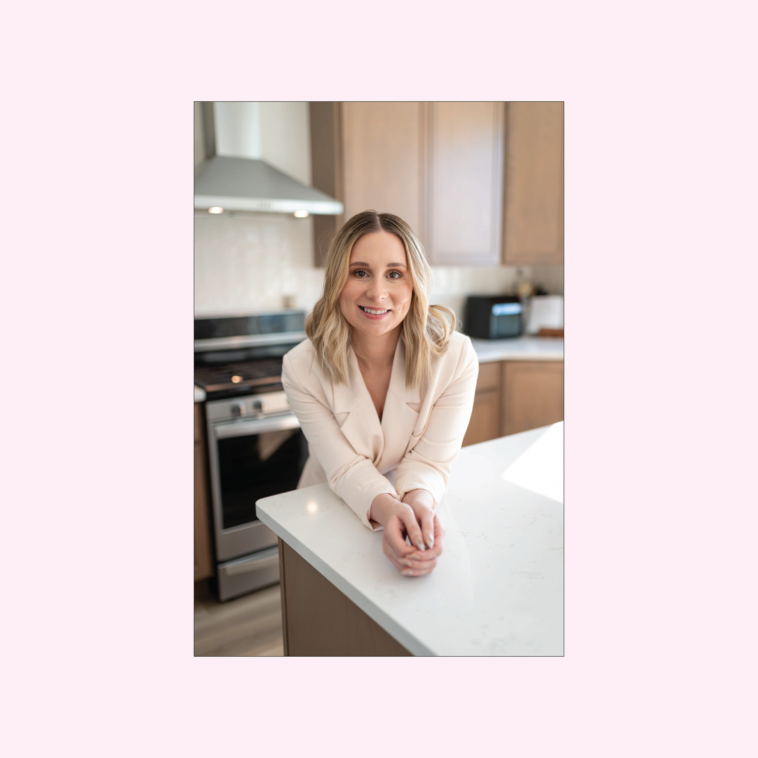 A smiling woman with blonde hair, wearing a beige blazer, leaning on a white kitchen counter in a modern kitchen. Kitchen appliances and cabinets are visible in the background.