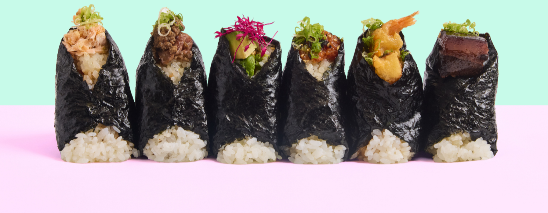Six onigiri handrolls arranged in a row on a pastel pink and green background, each wrapped in seaweed and filled with rice and various toppings.