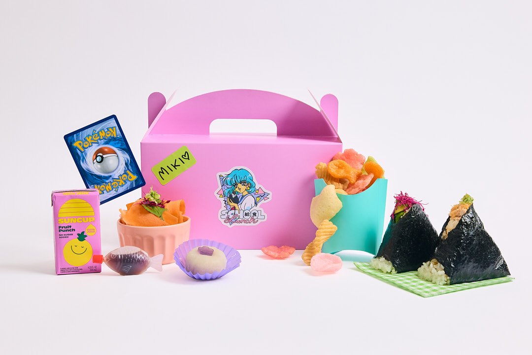 A modern bento box including a pink box, candy, snacks, suncup fruit juice box, a Pokémon card, and onigiri rice balls, all arranged on a white surface. 