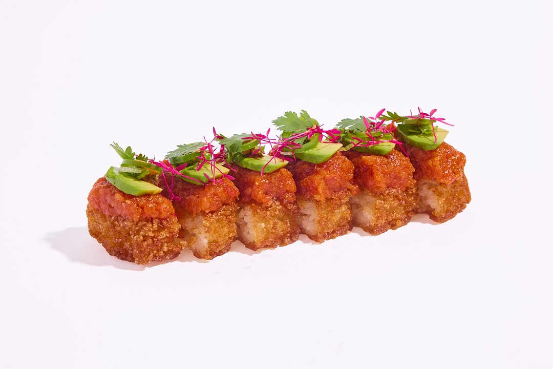 Six pieces of spicy tuna crispy ricecake topped with avocado slices, pink microgreens, and a spicy mayo against a white background.