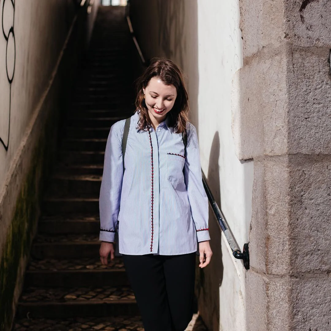 A young woman with shoulder-length brown hair, wearing a light blue striped shirt and black pants, smiling as she walks down a narrow outdoor staircase.
