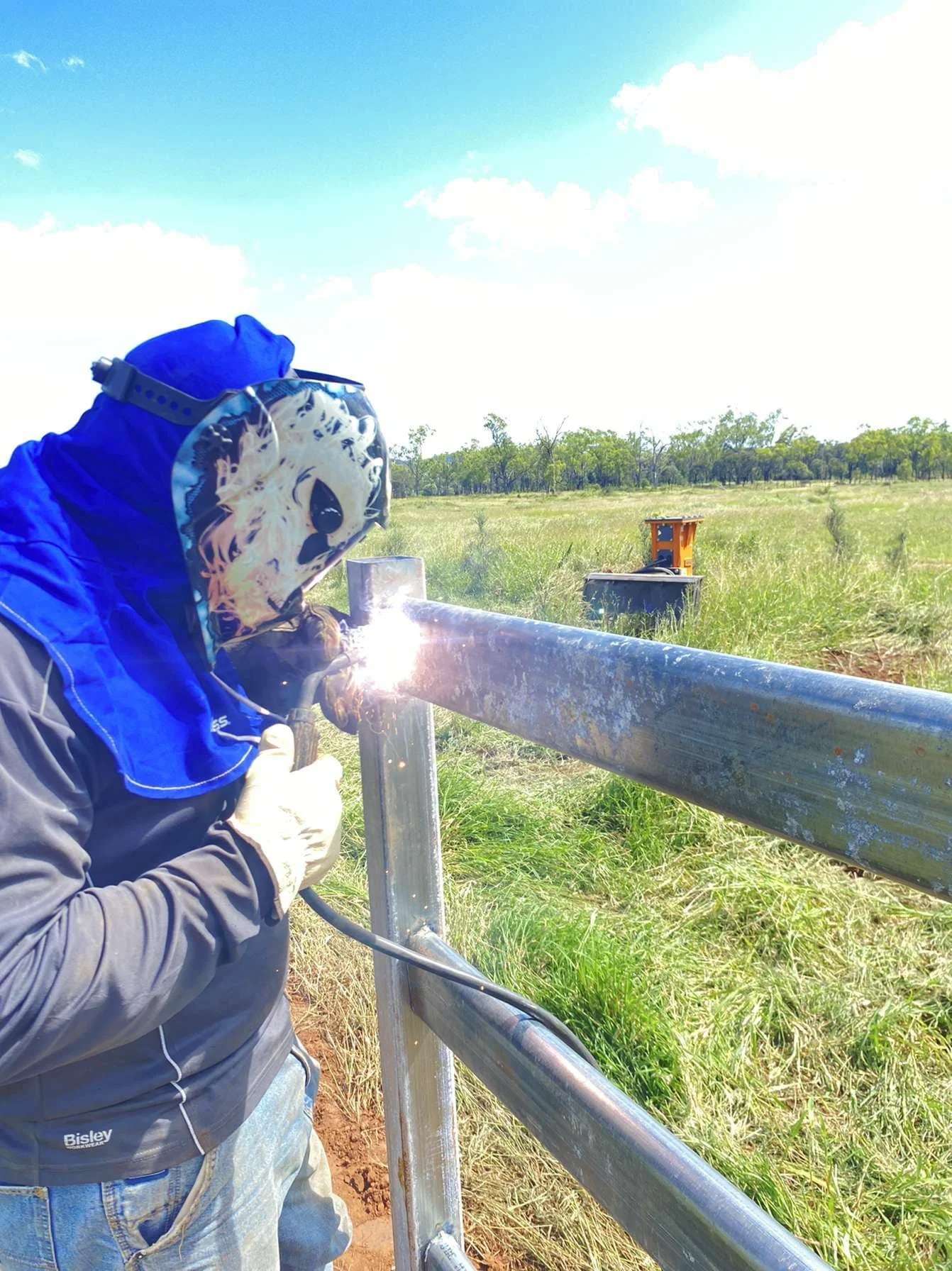 A person welding a metal gate outdoors on a sunny day, wearing protective gear including a welding helmet and gloves, in a grassy field.