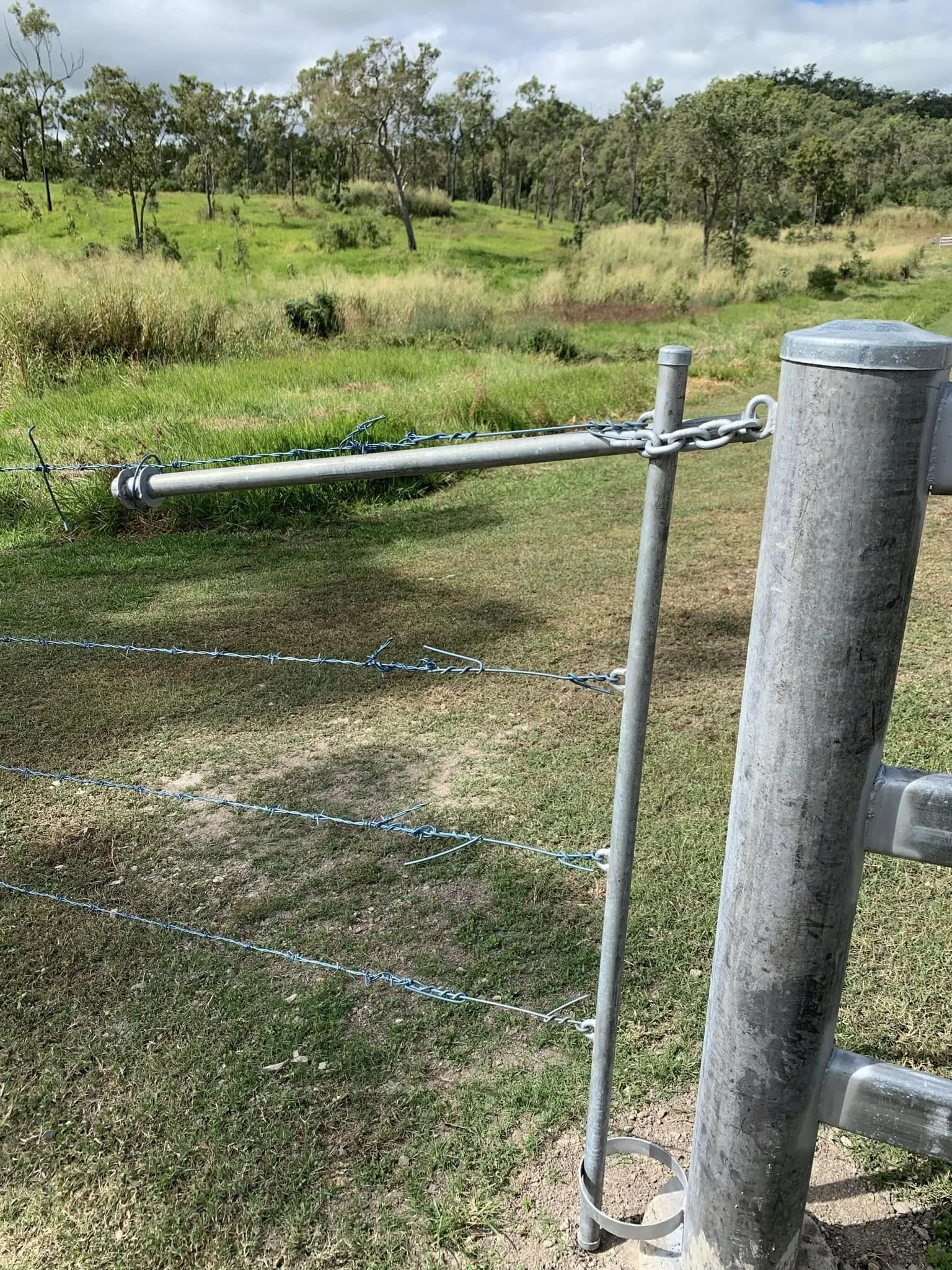 A metal fence with barbed wire in a grassy field with trees and hills in the background.