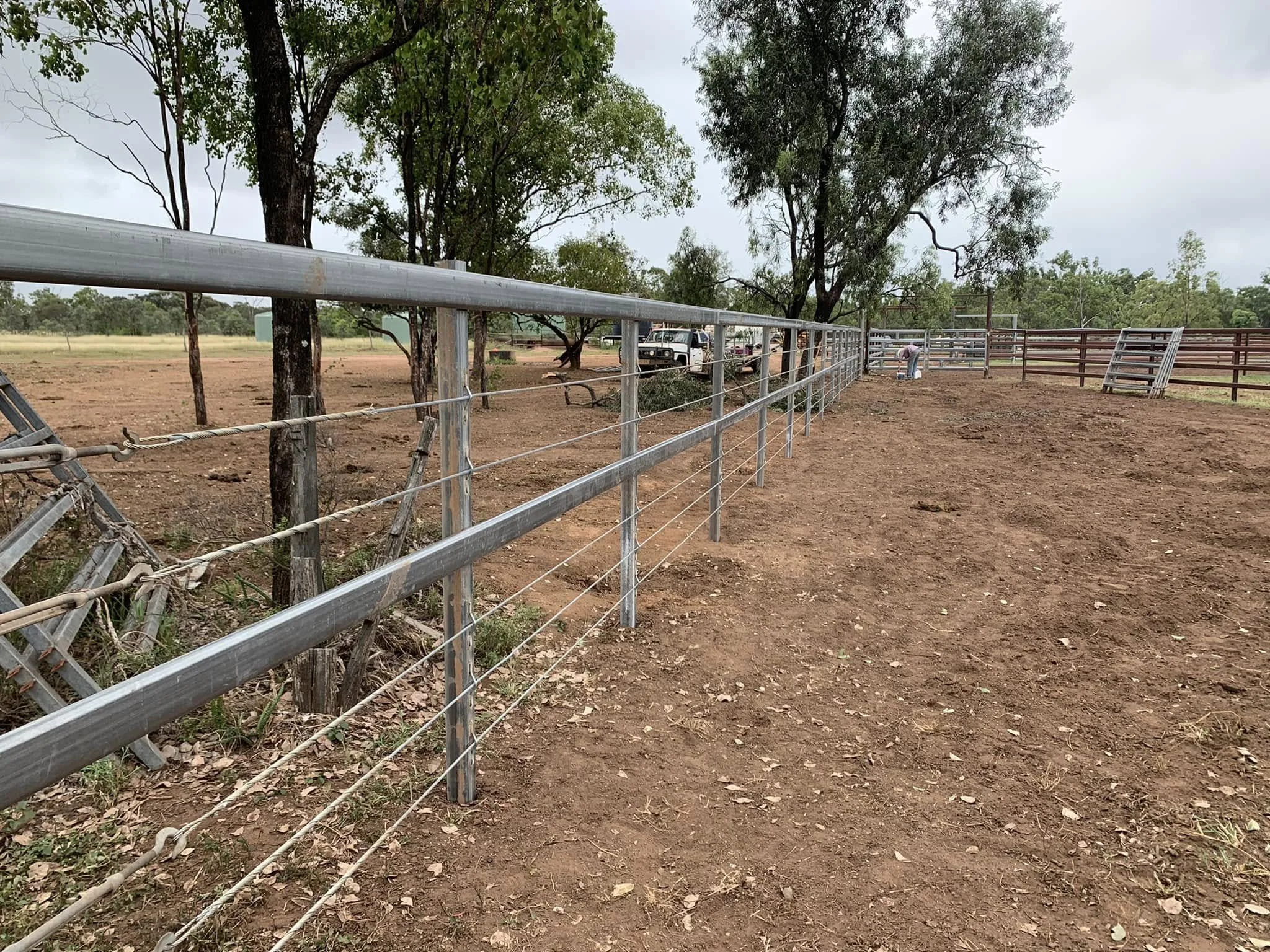 View of a fenced outdoor area with trees, dirt ground, and some equipment, possibly a ranch or farm setting.