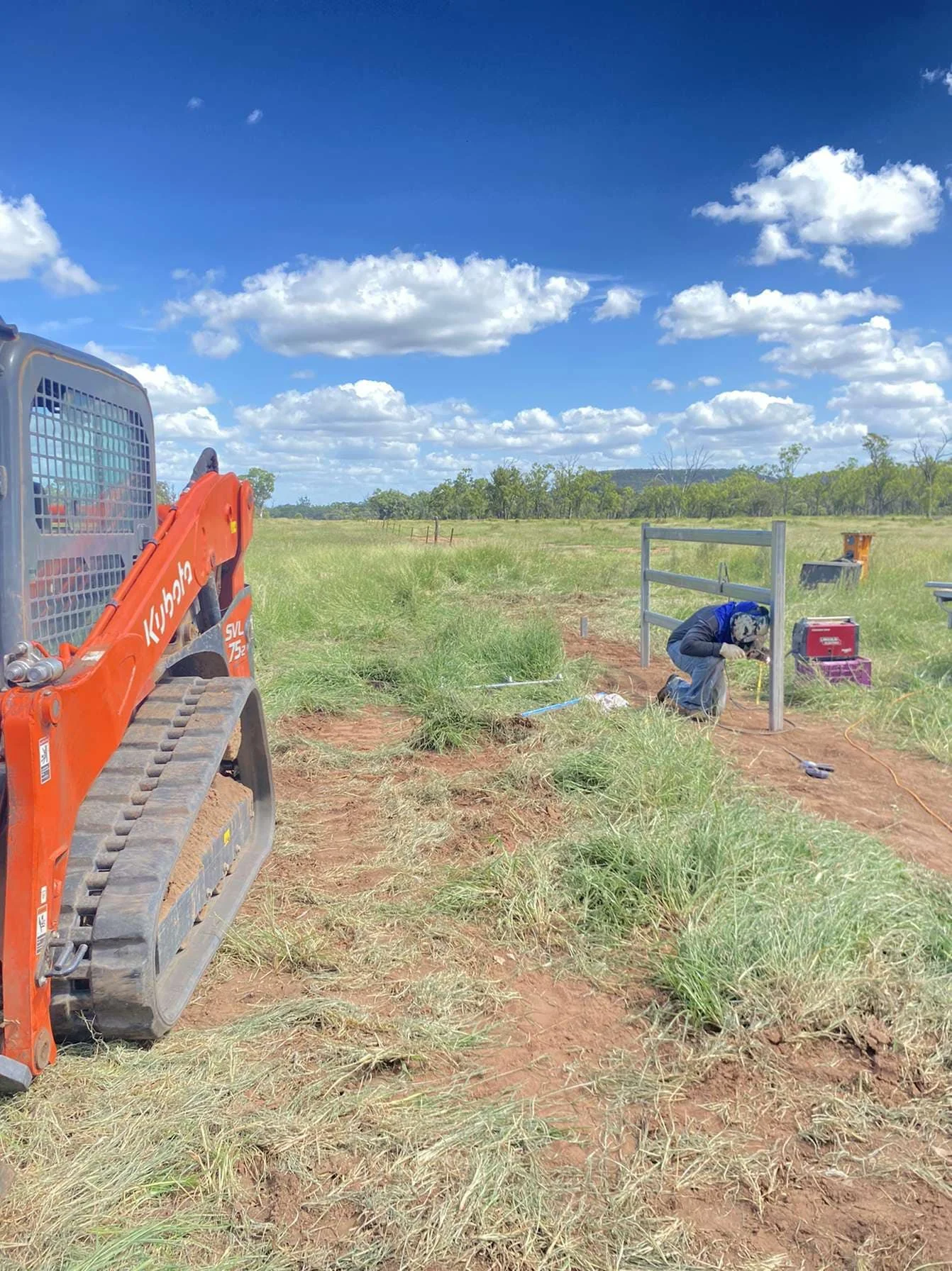 A construction site in an open field with a person welding a metal fence panel, a small orange Kubota compact excavator on the left, and a clear blue sky with scattered clouds.