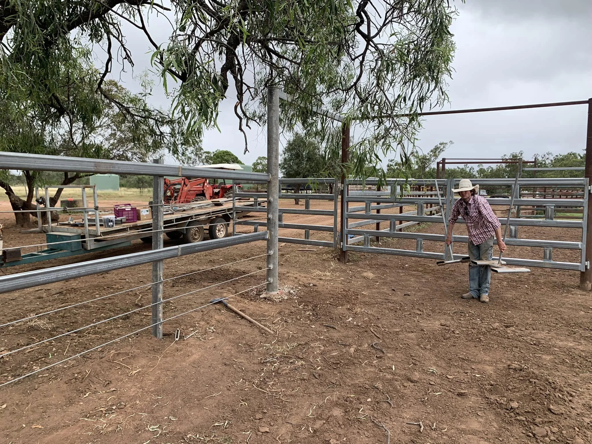 A woman wearing a cowboy hat and plaid shirt is installing a metal gate on a dirt farm or ranch. There are metal fences, a truck with tools and equipment, trees, and open land in the background.