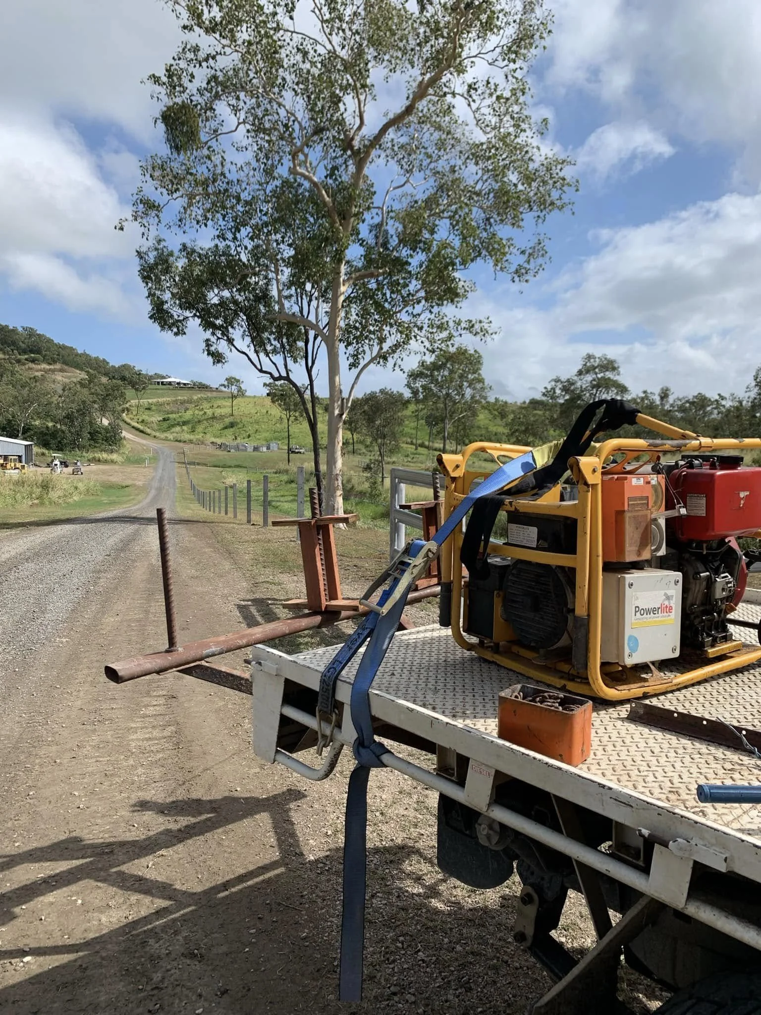 A dirt road running through a rural landscape with green hills, trees, and a partly cloudy sky. A utility cart with construction equipment is parked on the side of the road.