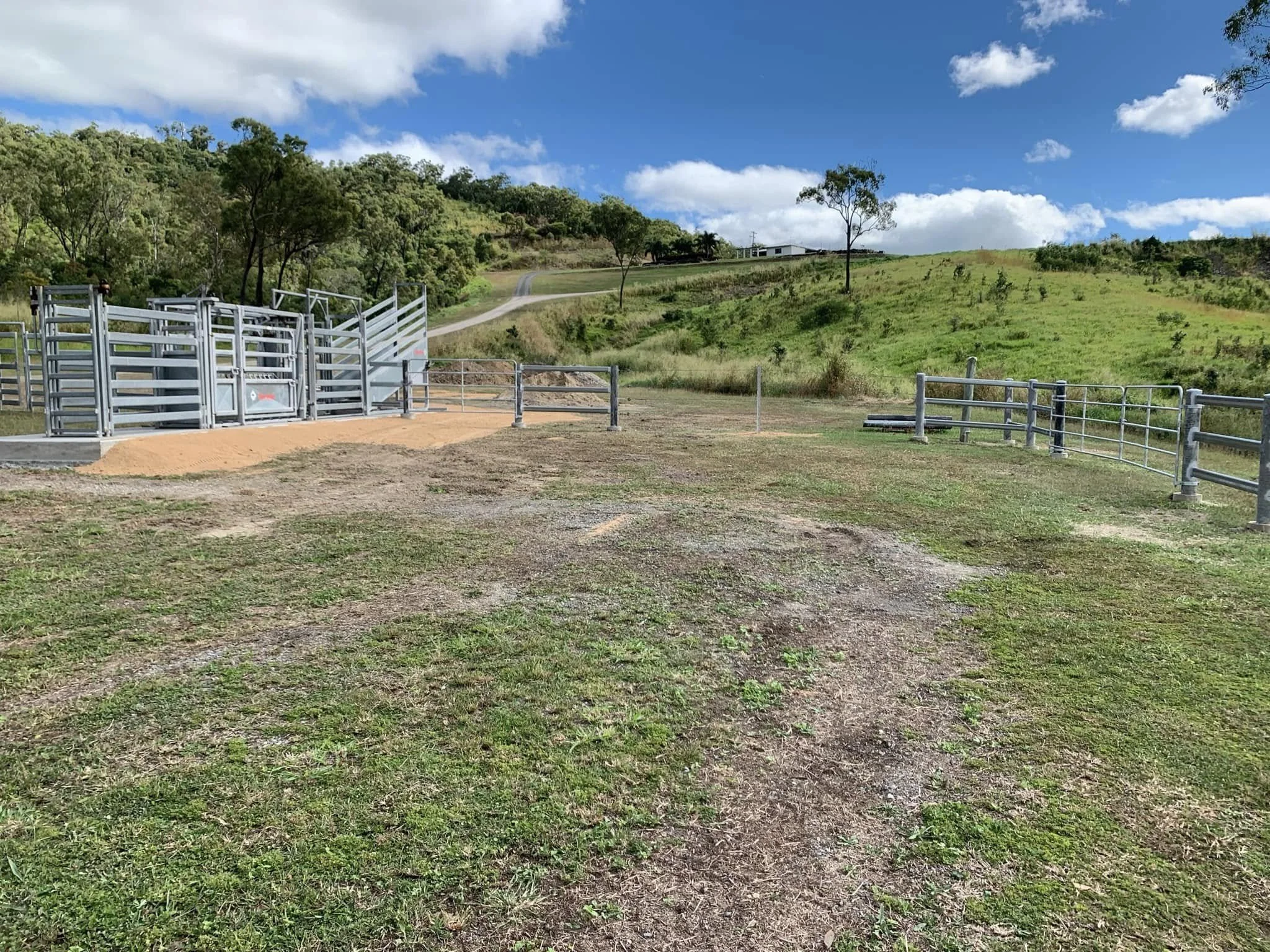 Open cattle gate on grassy hillside with green trees, blue sky, and scattered clouds in the background.
