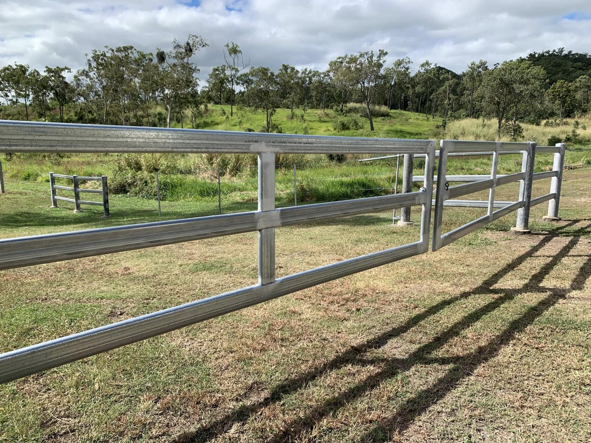 A metal fence with horizontal bars and vertical posts on a grassy field with trees and a cloudy sky in the background.