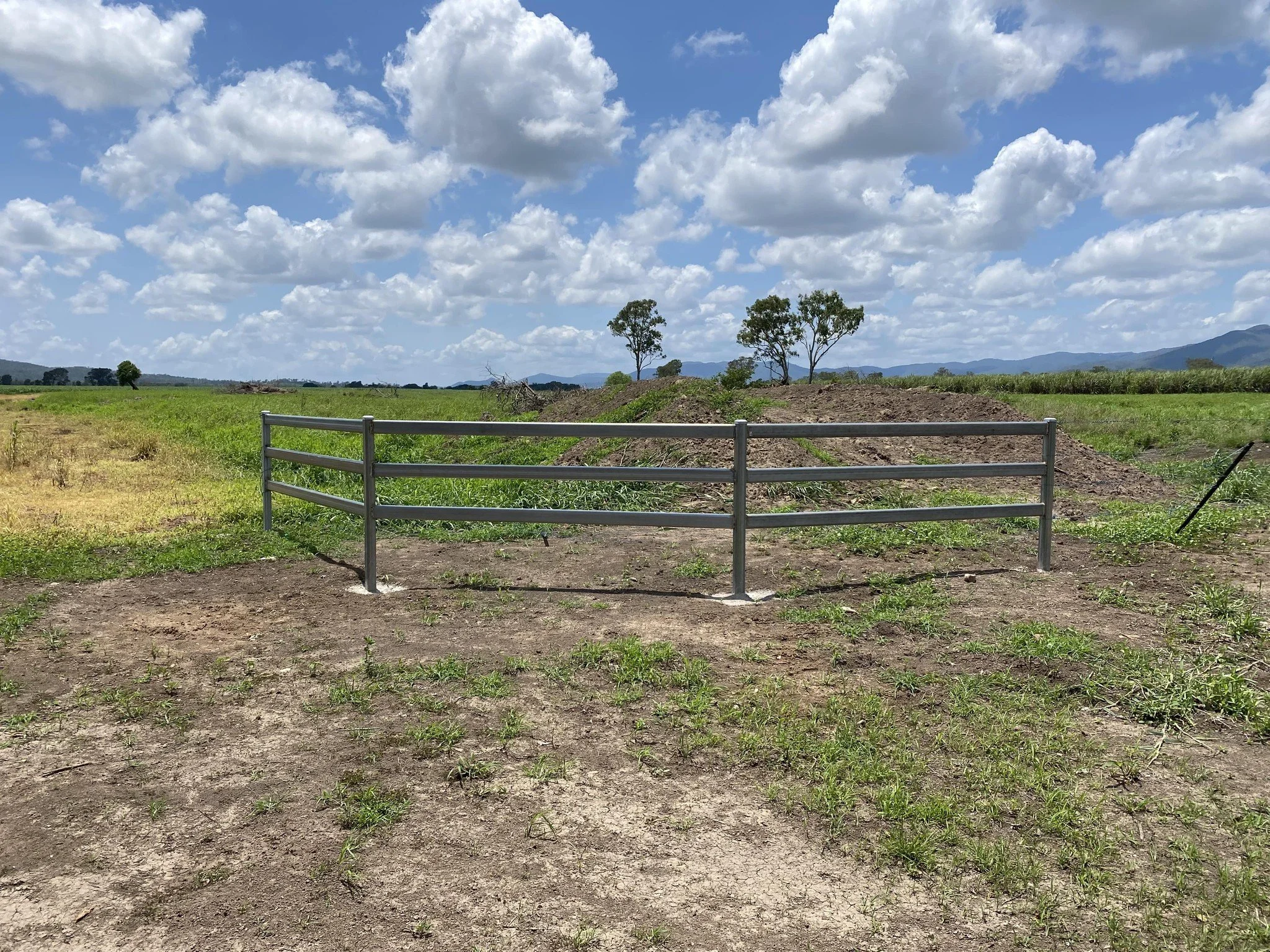 A metal fence post with three horizontal bars in an open field with green grass, trees, distant hills, and a partly cloudy blue sky.