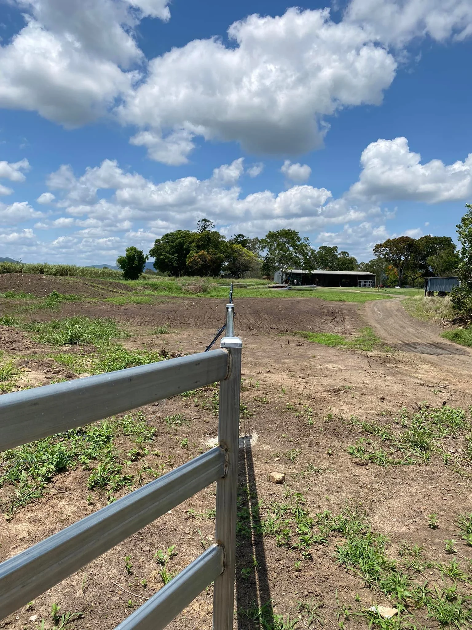A rural farm scene with a dirt road and metal fencing, green fields, trees, farm buildings, and a partly cloudy sky.