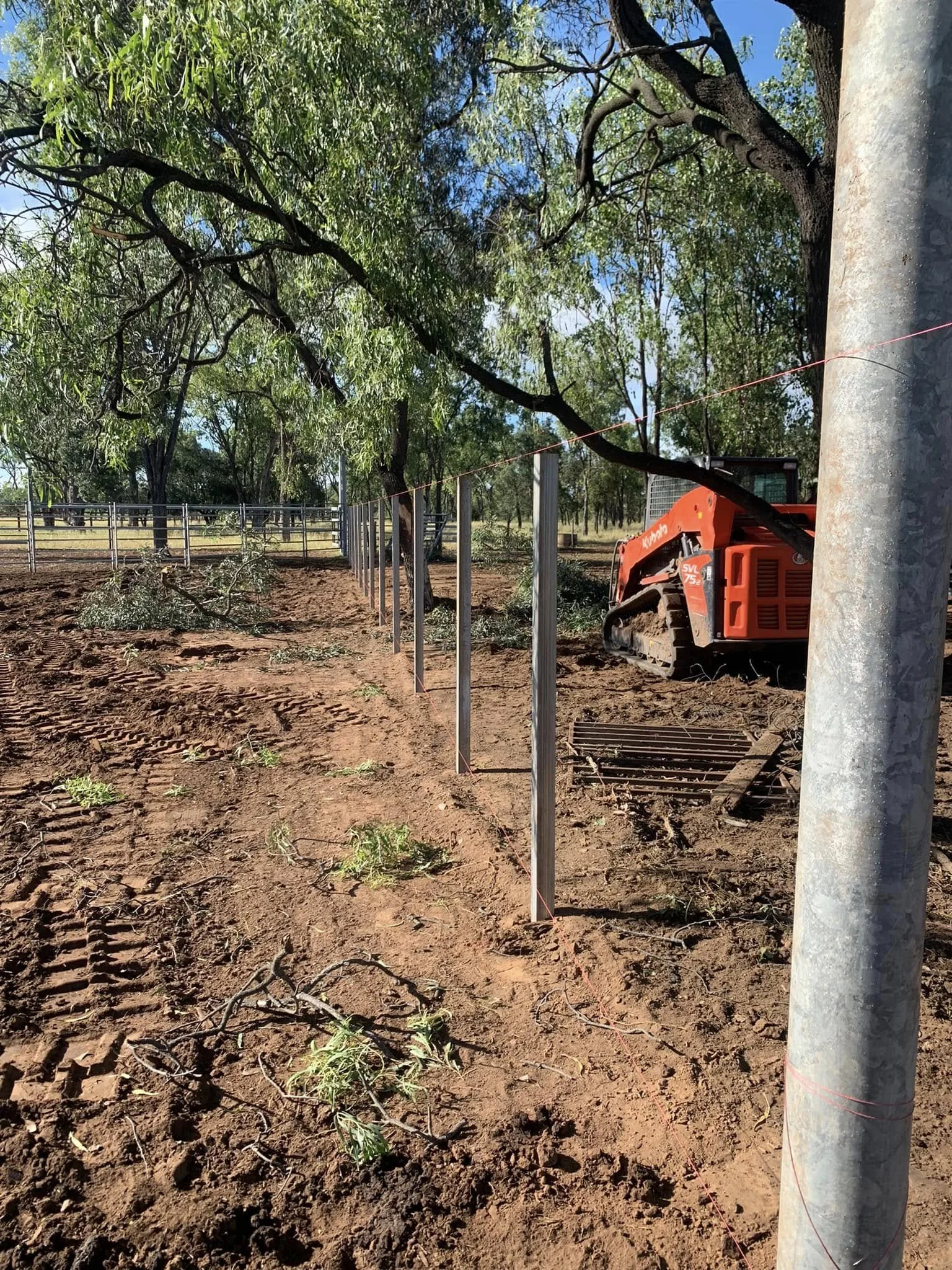 Construction site with a red excavator behind a metal fence, trees in the background, and dirt ground with tire tracks and scattered branches.