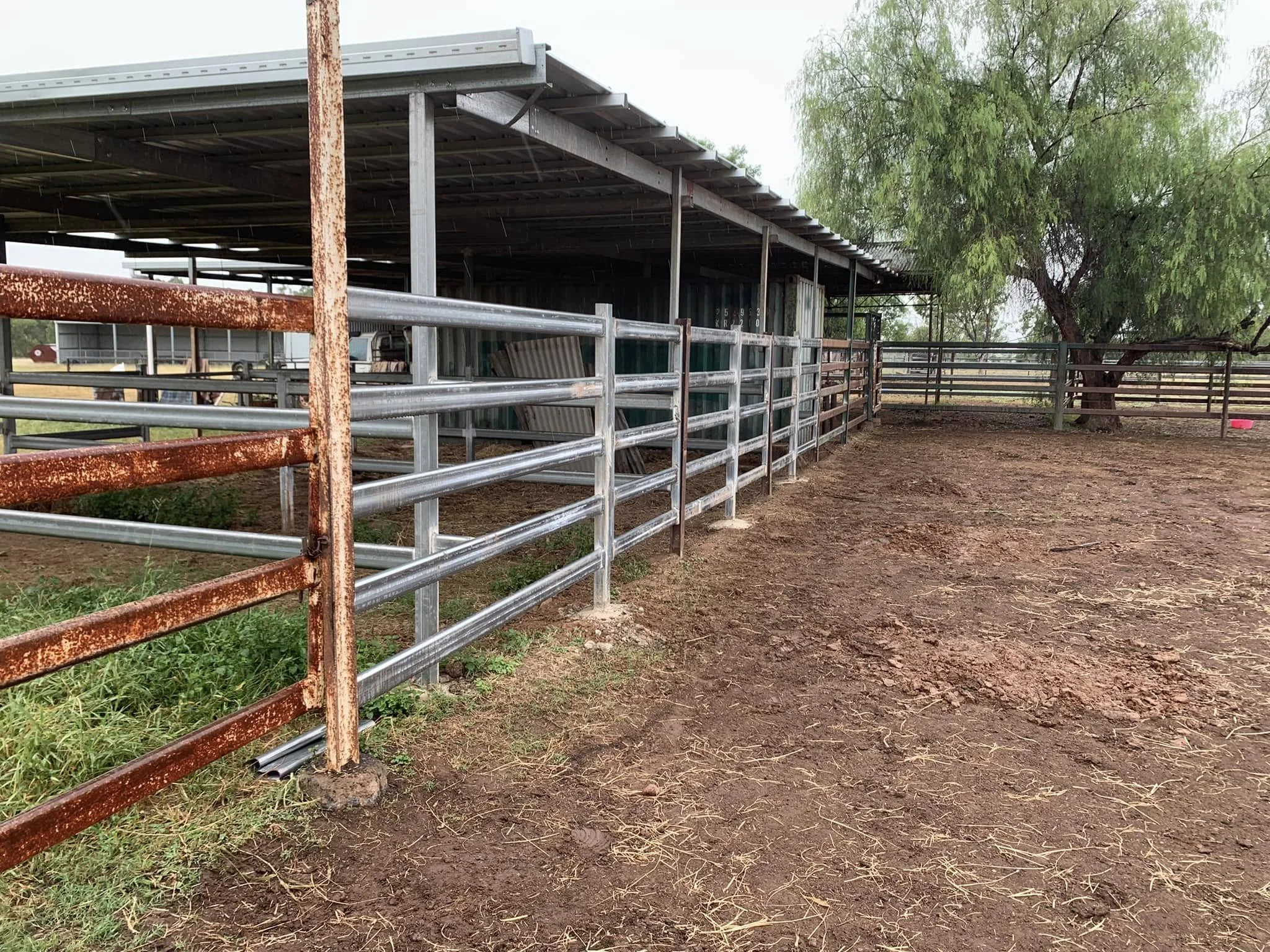 A fenced area at a farm with a metal shelter, some rusted and new metal fences, a tree, and a dirt ground.