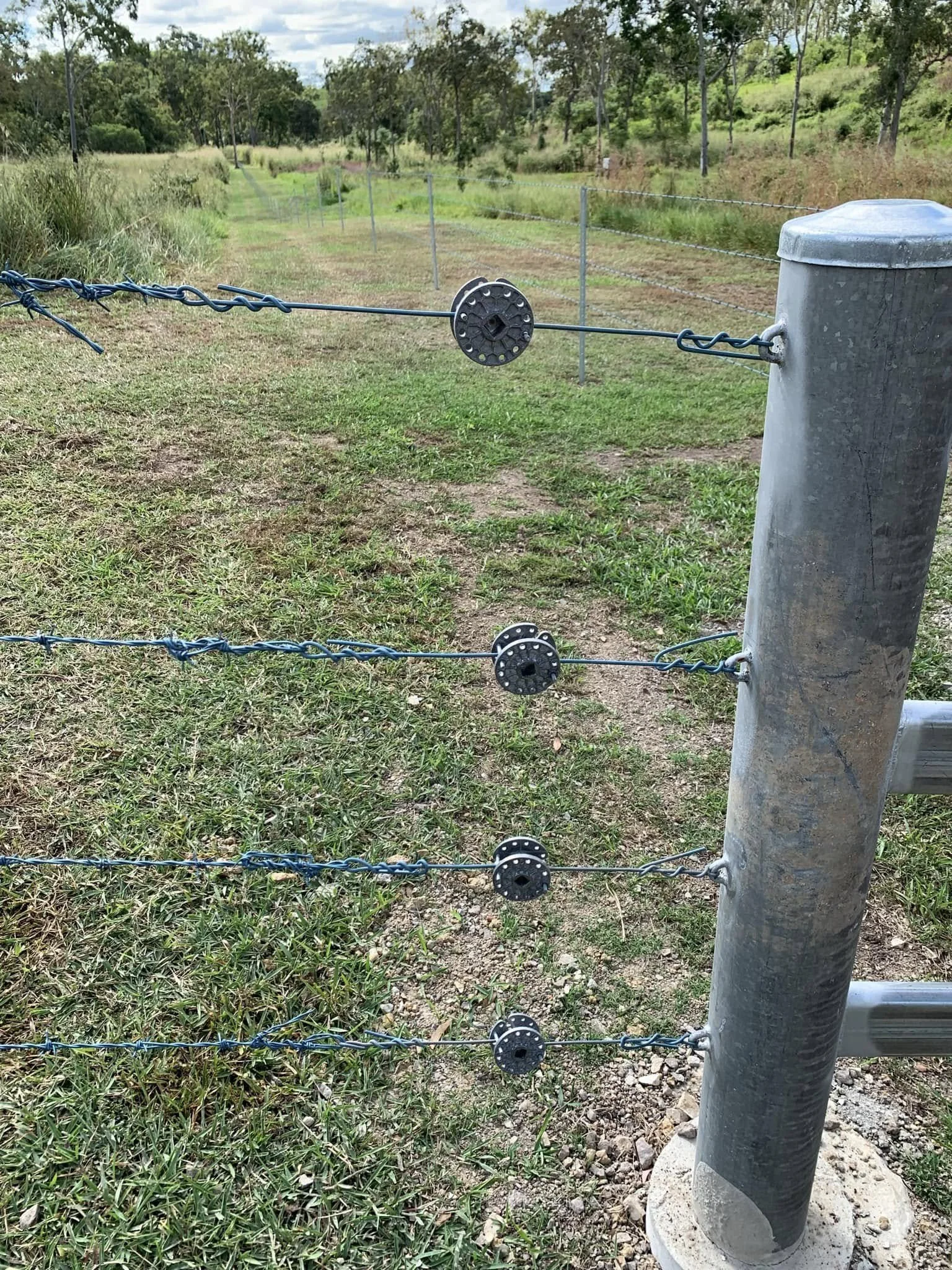 Barbed wire fence with three strands, supported by a metal post in an outdoor grassy area with trees and a clear sky in the background.