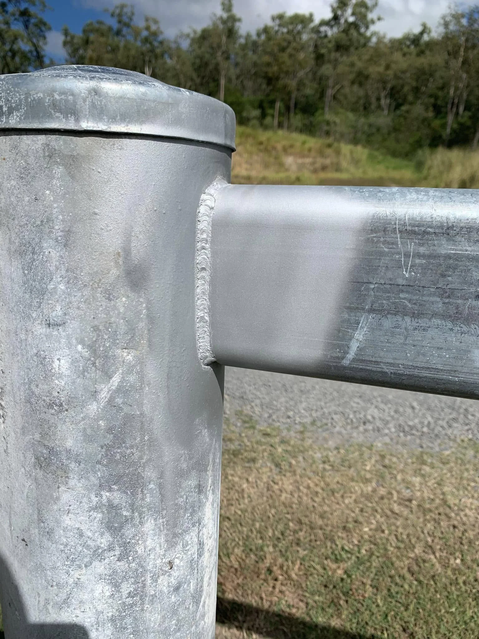 Close-up of a galvanized metal fence post with a horizontal metal rail attached, outdoors with a background of trees and grassy area.