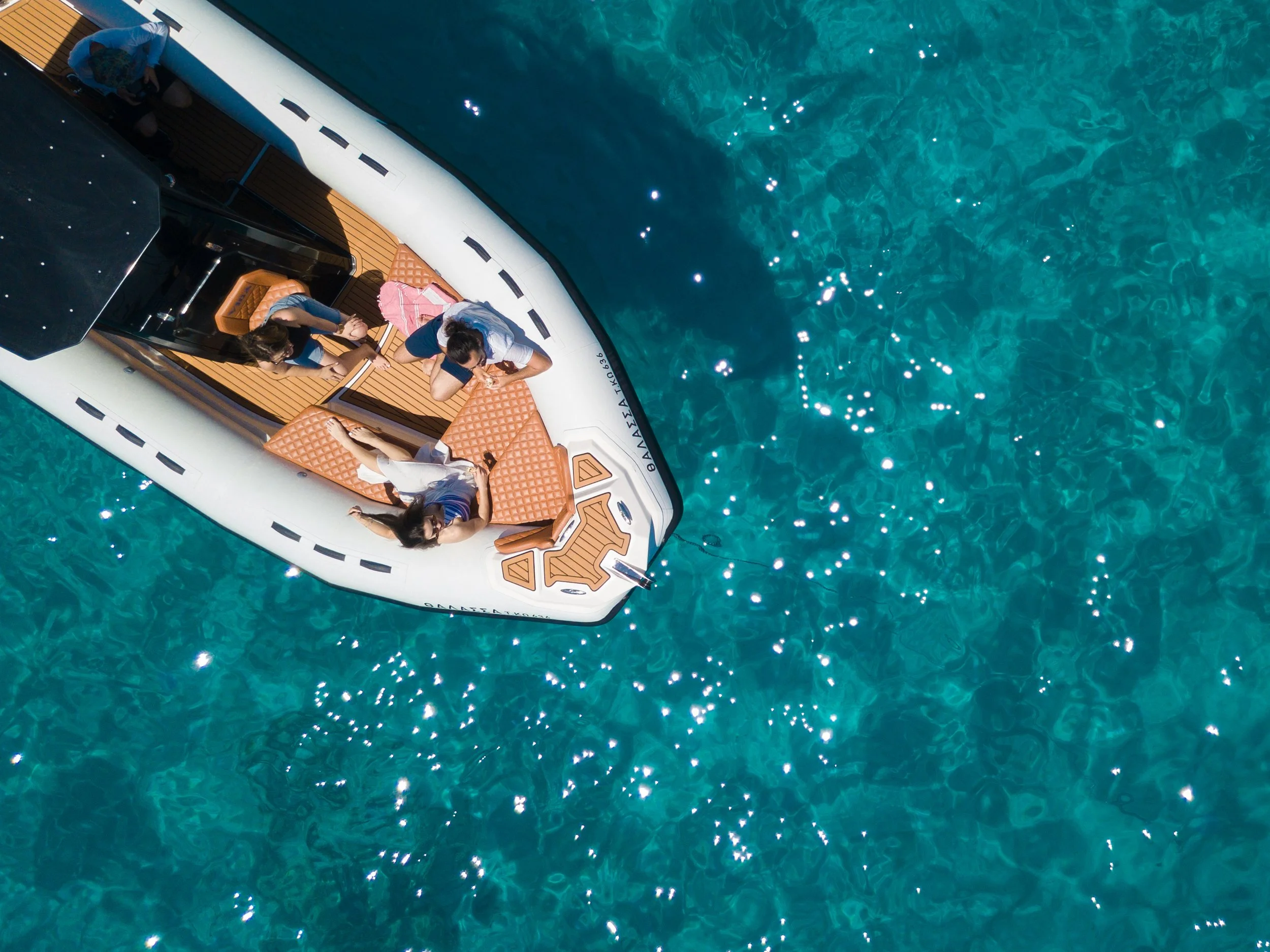 People relaxing on a yacht with wooden flooring, floating on clear blue water, some sitting, some lying down, with a pink towel and personal belongings nearby.